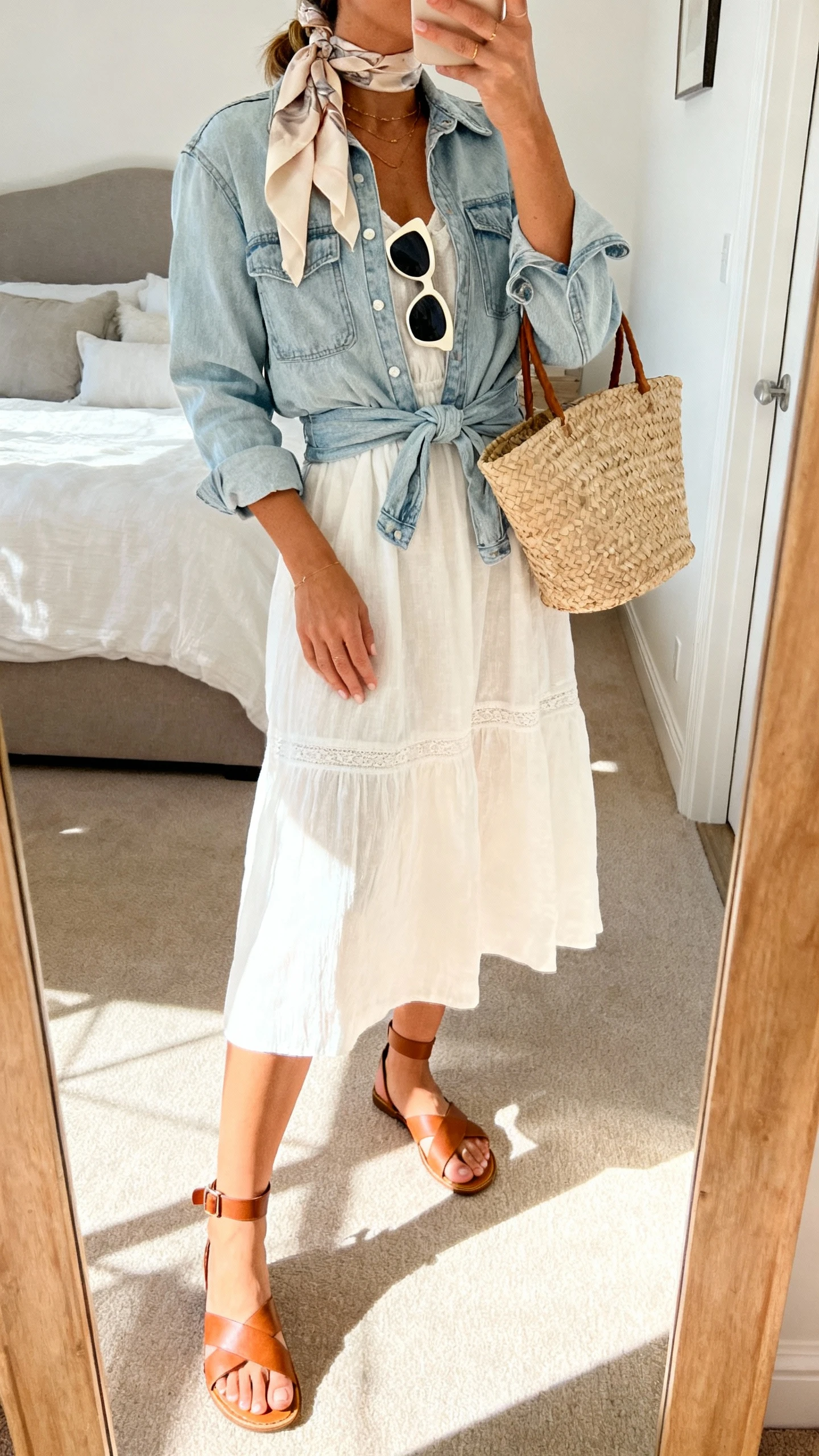 iPhone mirror selfie of a woman wearing a coastal gelato stroll look: airy cotton sundress, light denim jacket tied at waist, comfy leather sandals with ankle strap, woven straw tote, cat-eye sunglasses, and a silk hair scarf; face not visible, cropped at shoulders, bedroom mirror with bright daylight, iPhone photo quality.
