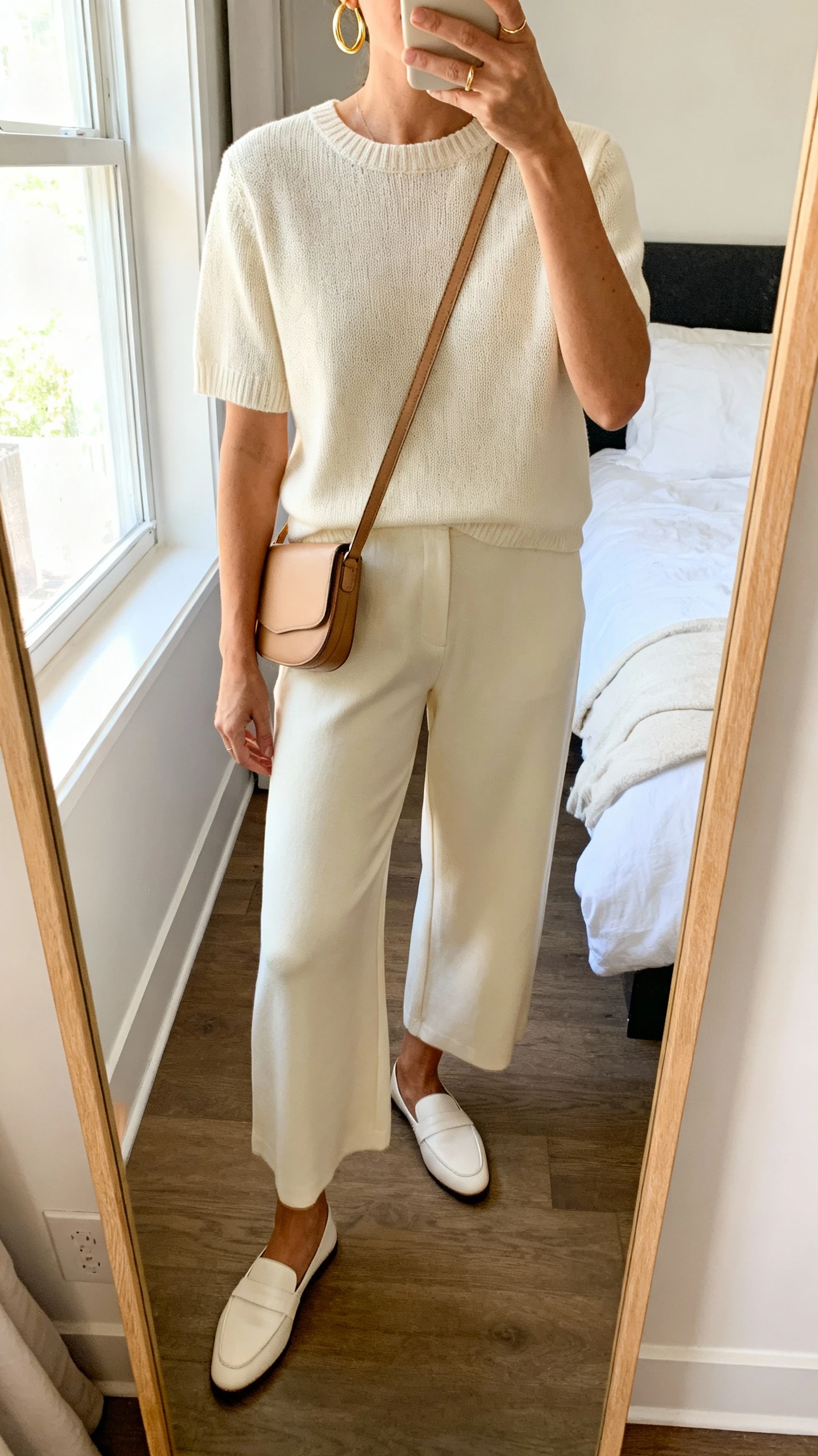 iPhone mirror selfie of a woman wearing a coordinated knit matching set (cream short-sleeve top and matching wide-leg pants) with minimalist gold hoops, a slim leather crossbody, and simple white loafers, face not visible, cropped at neck, bedroom mirror, morning window light, iPhone photo quality