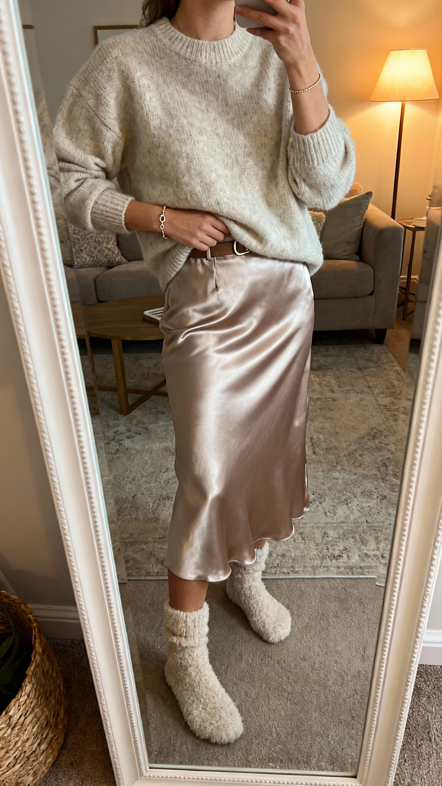 iPhone mirror selfie of a woman wearing a cozy cashmere sweater tucked into a satin midi skirt, fuzzy socks and a dainty bracelet, face not visible, cropped at neck, living room mirror with warm lamp light for a cozy vibe, iPhone photo quality