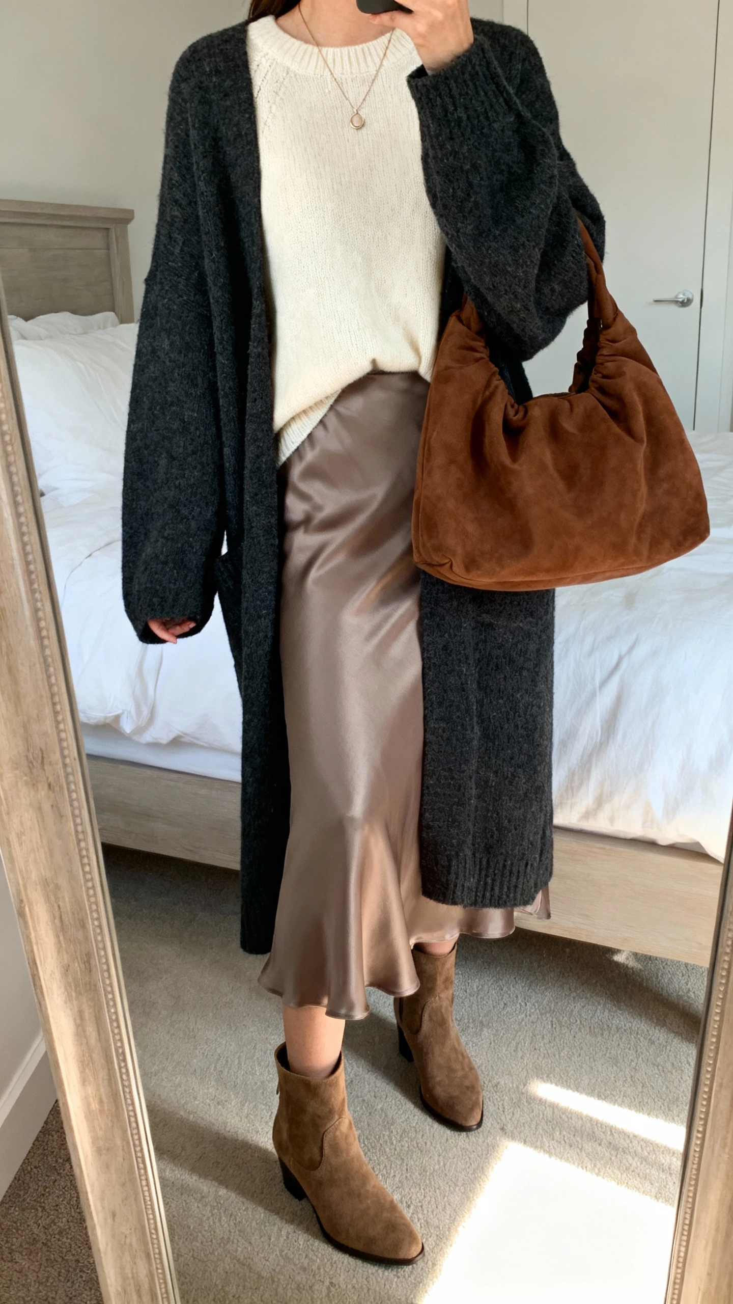iPhone mirror selfie of a woman wearing a cream cashmere sweater over a taupe satin slip skirt, oversized charcoal cardigan, suede ankle boots, slouchy chocolate hobo bag, dainty pendant necklace, face not visible, cropped at shoulders, bedroom mirror with soft afternoon light, iPhone photo quality.