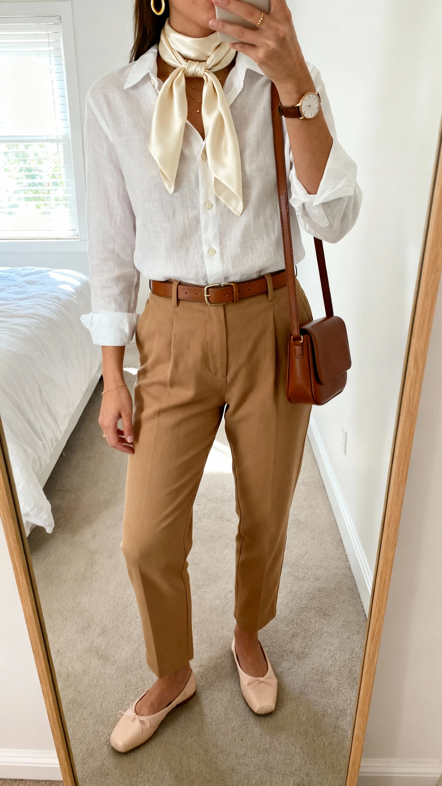 iPhone mirror selfie of a woman wearing a cream silk scarf tied around the neck, a lightweight white button-up tucked into high-waisted tan trousers, slim leather belt, ballet flats, structured brown crossbody, delicate gold hoops and watch, face not visible, cropped at neck, bedroom mirror with natural window light, iPhone photo quality