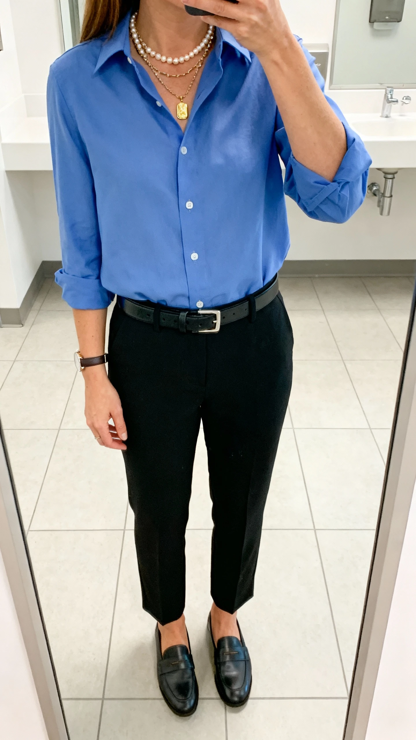 iPhone mirror selfie of a woman wearing a crisp blue button-down tucked into tailored black trousers, loafers, a structured belt, layered necklaces (pearl strand + gold pendant + subtle chain), and a slim watch, face not visible, cropped at shoulders, office bathroom mirror with bright indoor lighting, iPhone photo quality.