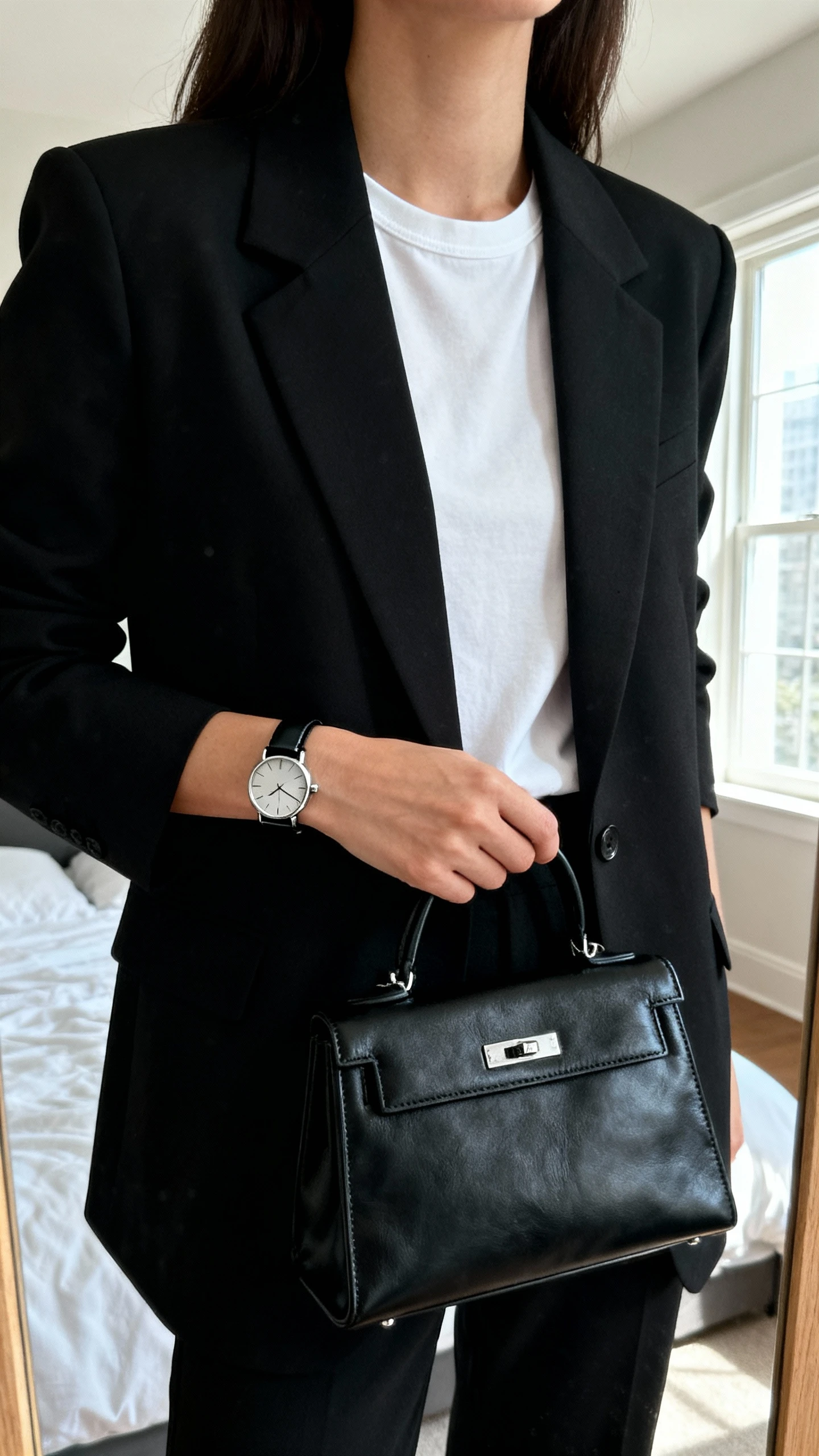 iPhone mirror selfie of a woman wearing a crisp monochrome city set: black tailored blazer, white crew-neck tee, black straight-leg trousers, sleek minimalist black leather watch with silver dial, black loafers, structured black leather handbag, face not visible, cropped at neck, bedroom mirror, natural window light, iPhone photo quality.