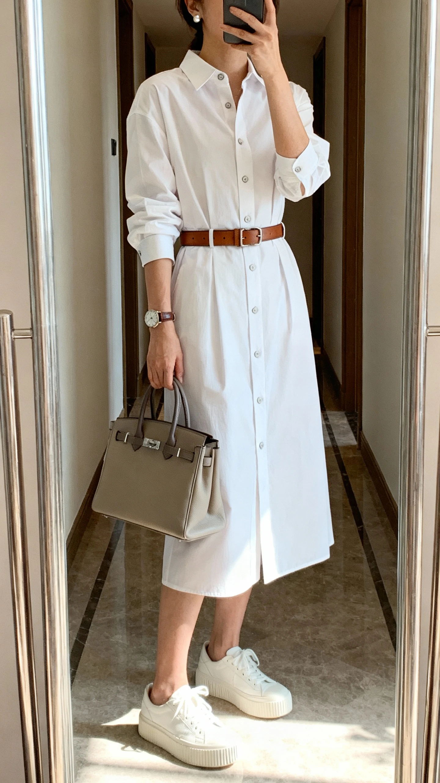 iPhone mirror selfie of a woman wearing a crisp shirt dress (button-front, tie-waist or straight cut), white platform sneakers, a structured tote or top-handle bag, a leather belt to define the waist (if no tie), a classic watch and stud earrings, face not visible, cropped at shoulders, full-length mirror in a hallway with indoor daylight, iPhone photo quality.
