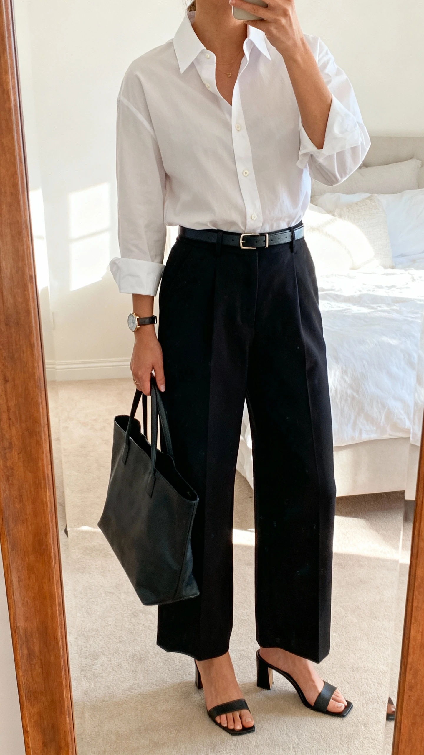 iPhone mirror selfie of a woman wearing a crisp shirt and wide-leg trousers: white button-up tucked into high-waist wide-leg black trousers, thin leather belt, heeled mules, understated watch, tote bag; face not visible, cropped at neck, bedroom mirror, bright morning light, iPhone photo quality.