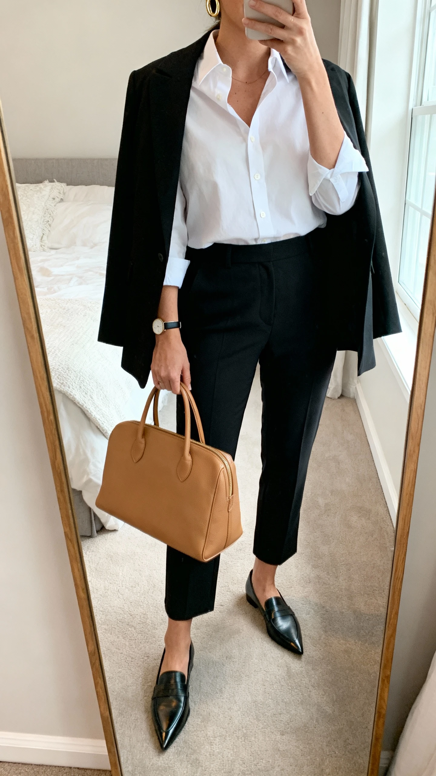 iPhone mirror selfie of a woman wearing a crisp white button-up tucked into tailored black ankle trousers, black blazer draped over shoulders, pointed-toe loafers, structured camel power tote, minimal gold hoop earrings and a slim watch, face not visible, cropped at neck, bedroom mirror, natural window light, iPhone photo quality.