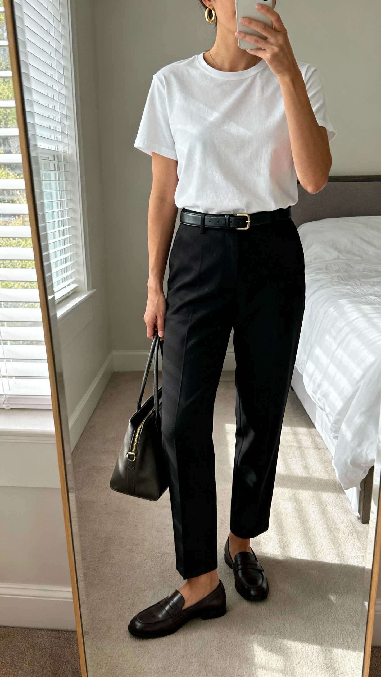 iPhone mirror selfie of a woman wearing a crisp white elevated tee tucked into tailored black trousers with a slim leather belt, minimal gold hoops, classic loafers, and a structured tote, face not visible, cropped at neck, bedroom mirror, soft morning window light, iPhone photo quality.