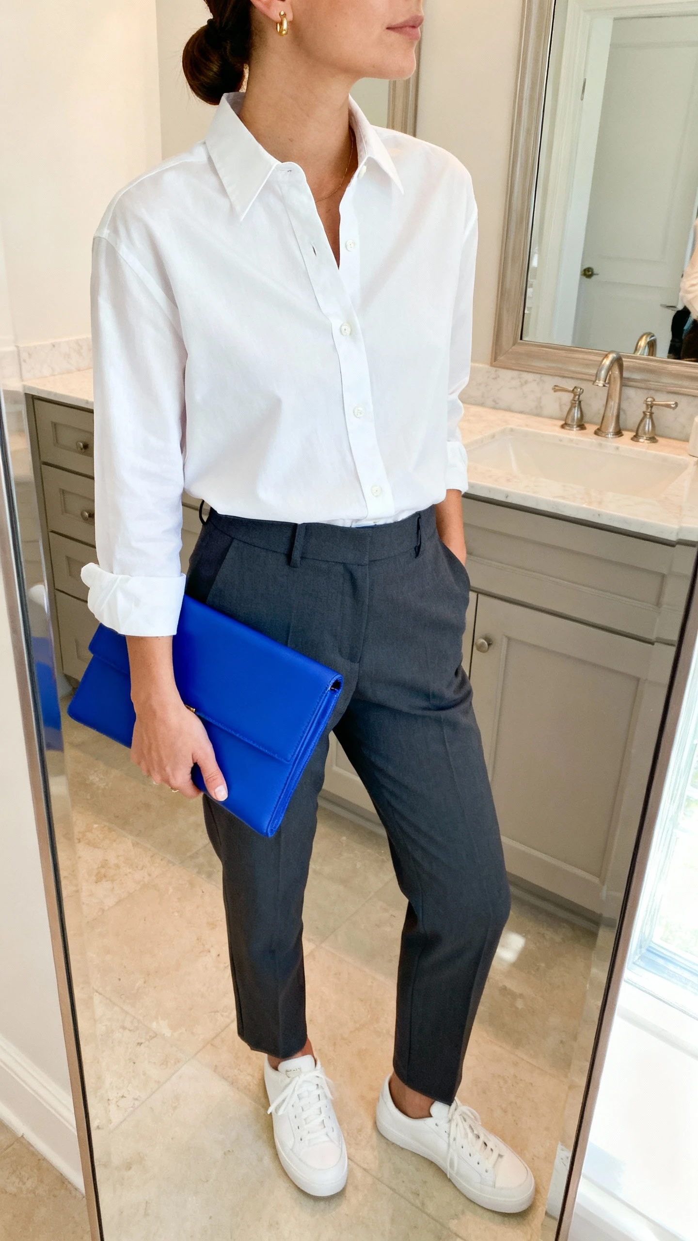 iPhone mirror selfie of a woman wearing a crisp white button-up tucked into tailored charcoal trousers, clean white sneakers, minimalist gold studs, slicked-back low bun, and carrying a bright cobalt structured clutch for a pop of color, face not visible, cropped at neck, bathroom mirror with soft natural light, iPhone photo quality.