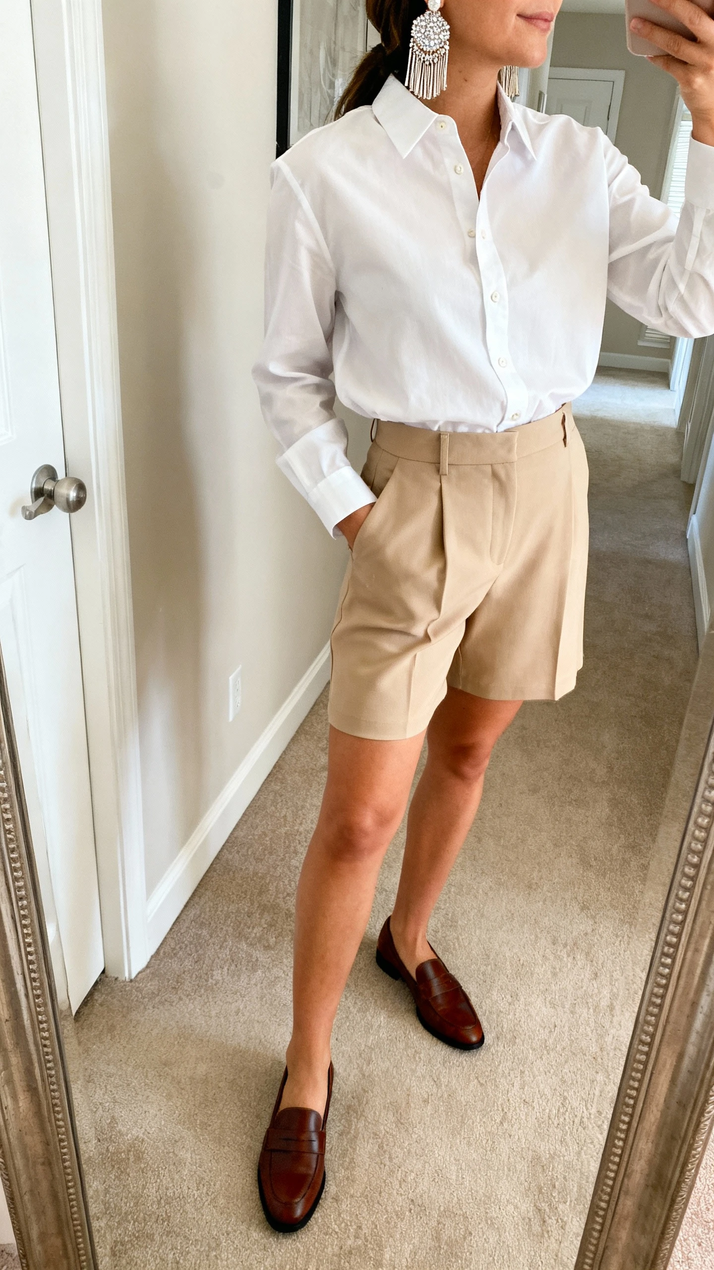 iPhone mirror selfie of a woman wearing a crisp white button-down tucked into tailored beige shorts, slim leather loafers, and glam fringe statement earrings with sparkle, face not visible, side angle in a hallway full-length mirror, natural morning light, iPhone photo quality.