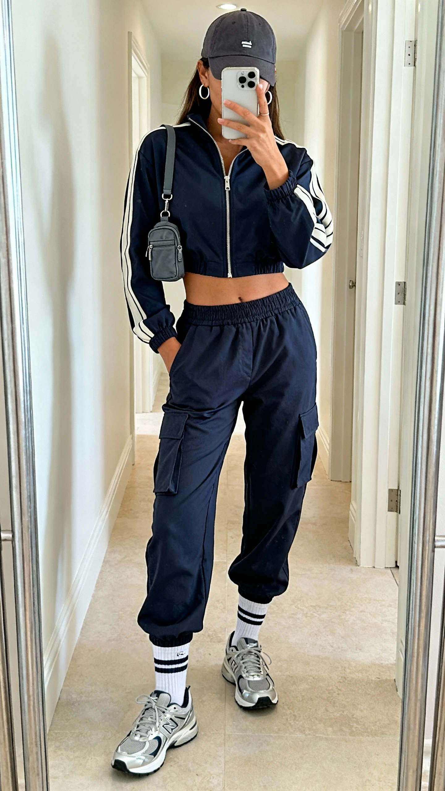 iPhone mirror selfie of a woman wearing a cropped zip-up track jacket, high-waisted cargo joggers, sleek trainers, sporty crew socks, a baseball cap, mini nylon crossbody, and subtle hoop earrings, face not visible, cropped at shoulders, hallway full-length mirror, bright indoor lighting, iPhone photo quality.