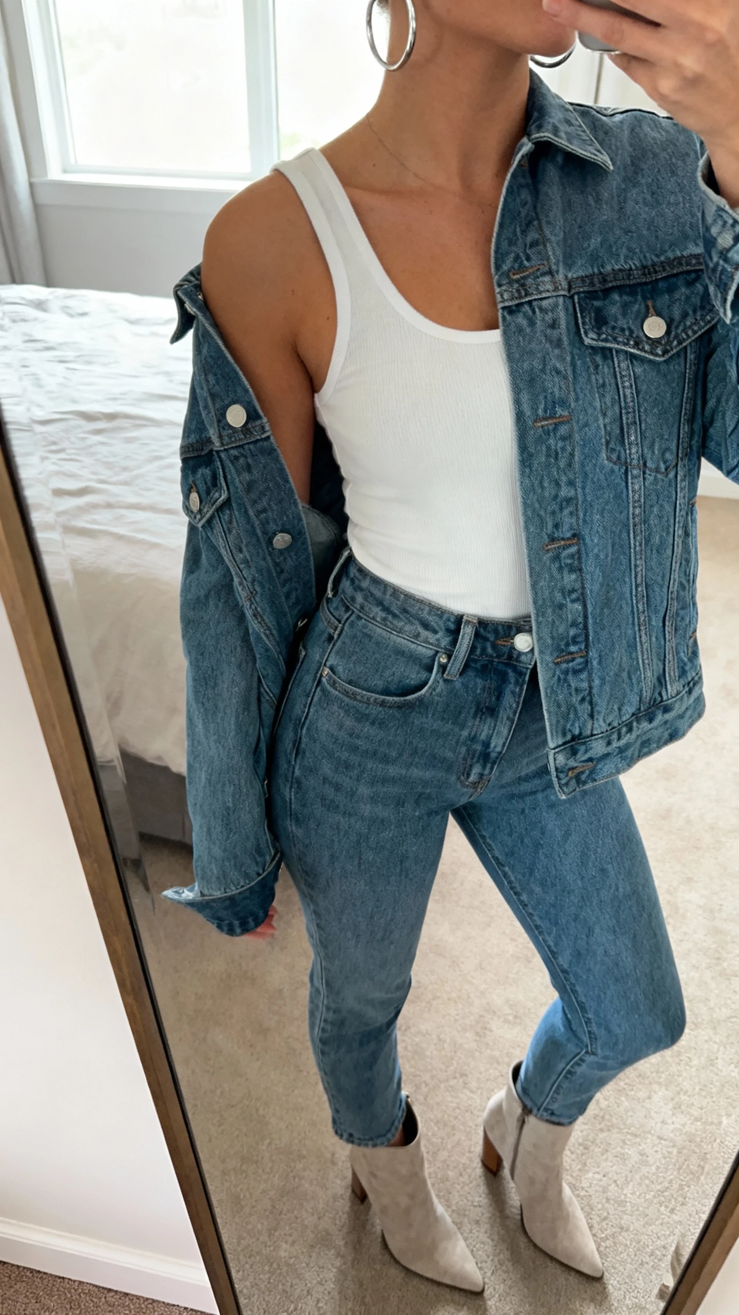 iPhone mirror selfie of a woman wearing a denim jacket with matching high-waisted denim jeans, a simple white tank underneath, heeled booties, and silver hoops, face not visible, cropped at shoulders, bedroom mirror, soft window light, iPhone photo quality.