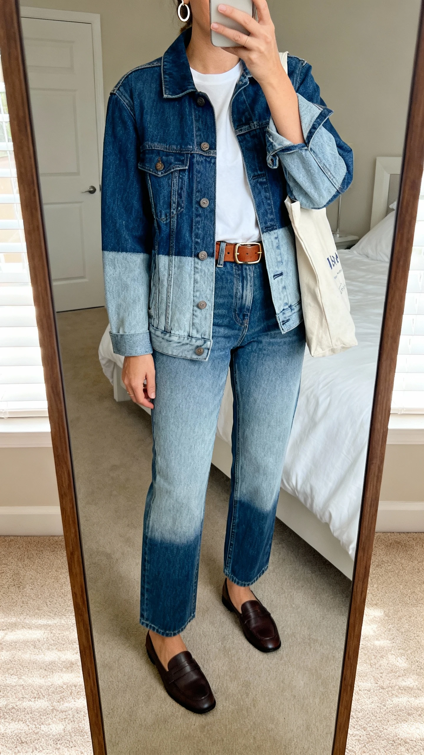 iPhone mirror selfie of a woman wearing a denim jacket with contrasting wash straight-leg jeans, white tee tucked in, leather belt, and loafers, subtle hoop earrings and a tote bag, face not visible, cropped at neck, bedroom mirror, natural daylight, iPhone photo quality