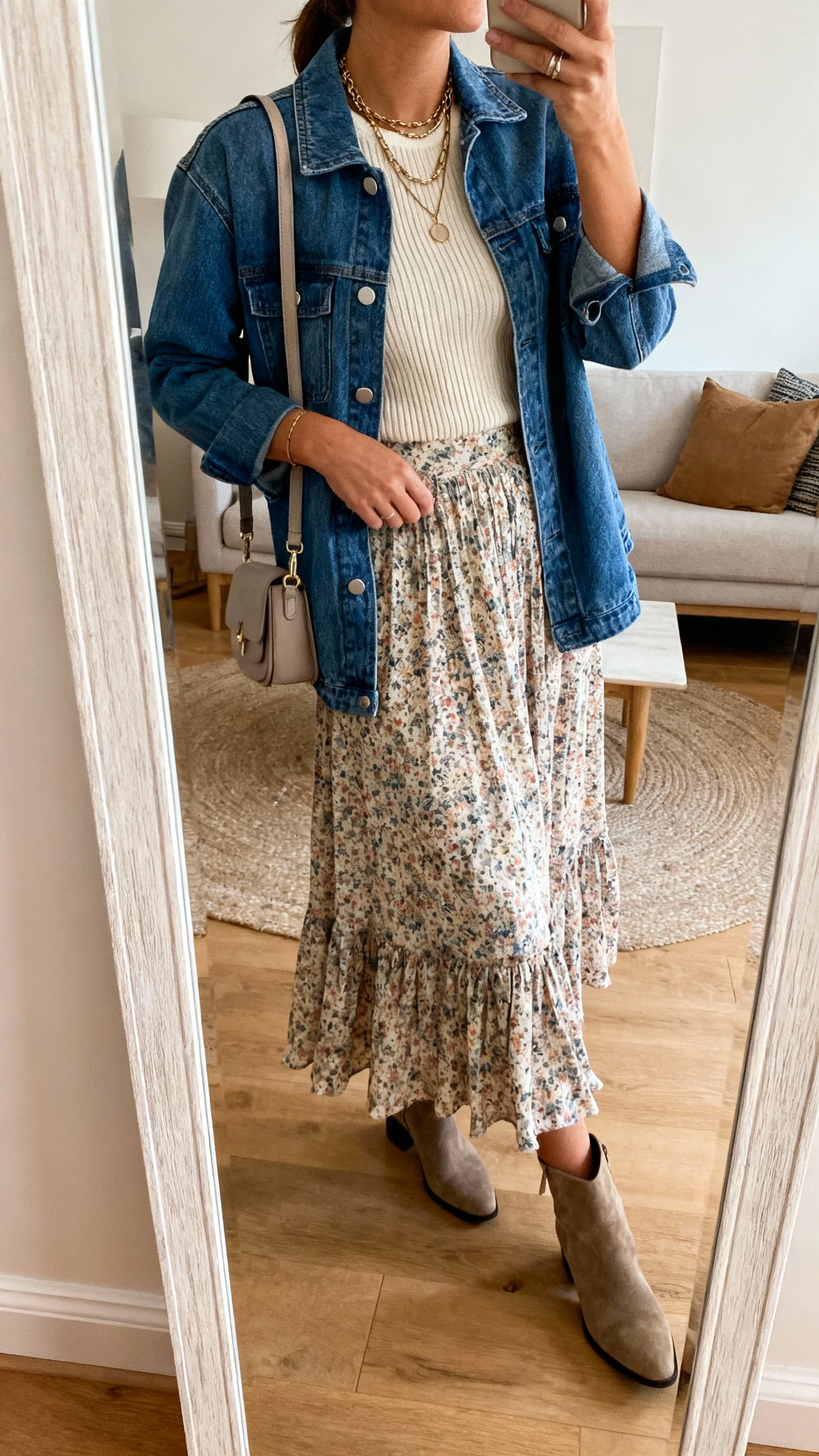 iPhone mirror selfie of a woman wearing a denim jacket over a flowy floral midi skirt, fitted knit top, ankle boots, layered necklaces, and a small shoulder bag, face not visible, side angle, living room full-length mirror, warm indoor lighting, iPhone photo quality