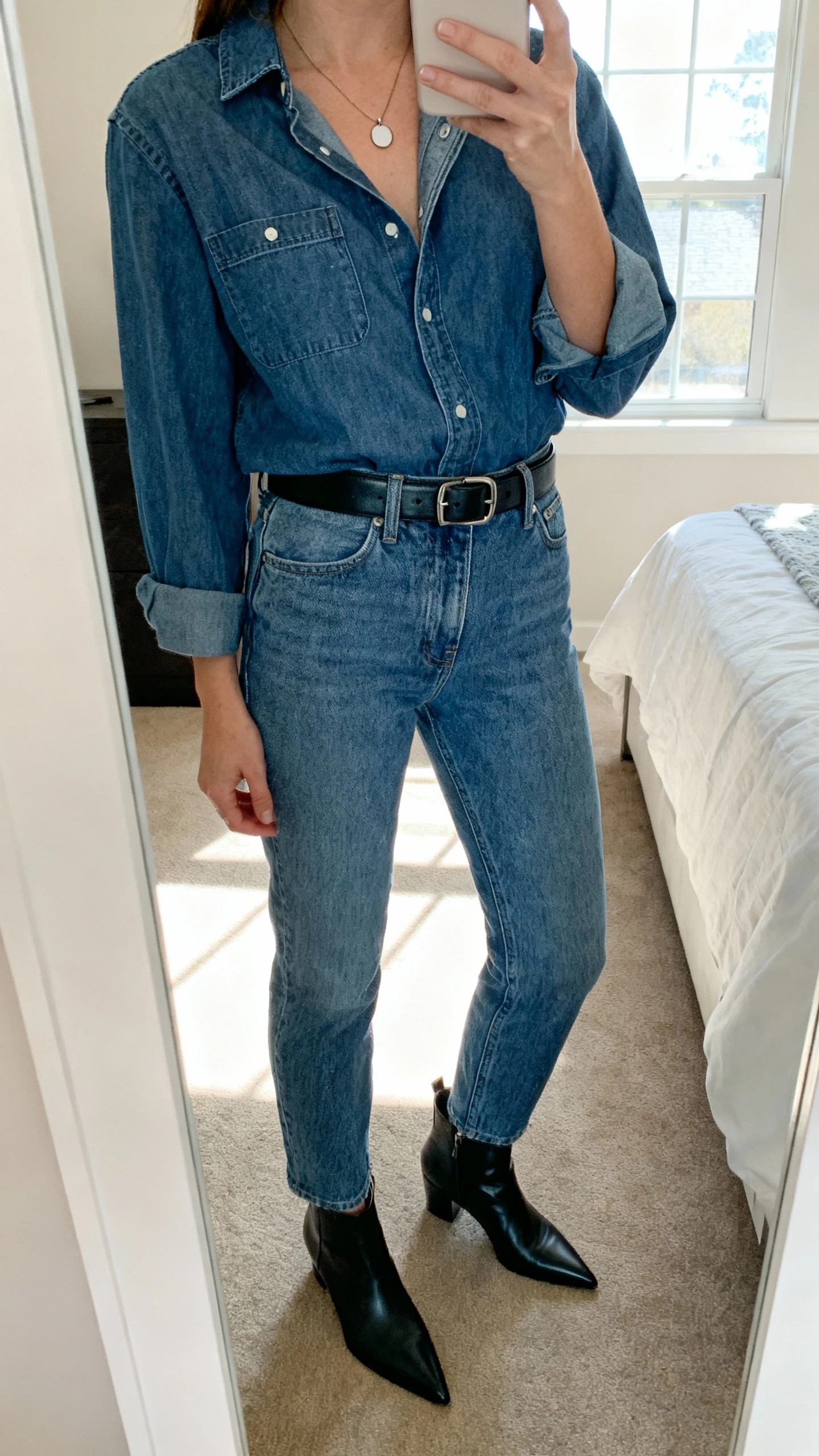 iPhone mirror selfie of a woman wearing a denim shirt tucked into matching high-waisted denim jeans, black leather belt, pointed ankle boots, and a simple pendant necklace, face not visible, cropped at neck, bedroom mirror with natural window light, iPhone photo quality.