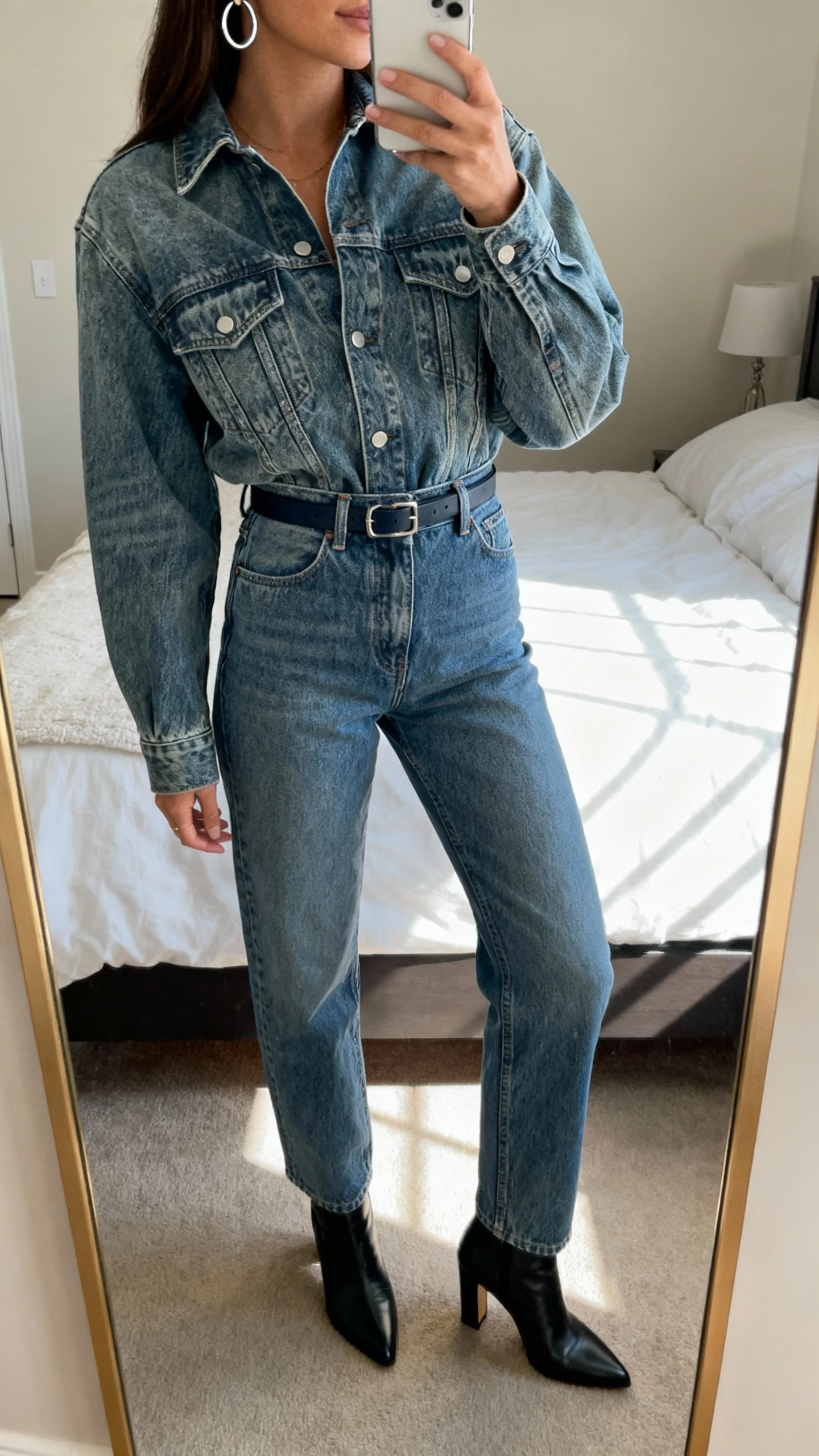 iPhone mirror selfie of a woman wearing a denim shirt jacket with vintage wash tucked into matching high-rise denim jeans, slim belt, heeled ankle boots, and silver hoop earrings, face not visible, cropped at shoulders, bedroom mirror, soft afternoon window light, iPhone photo quality.