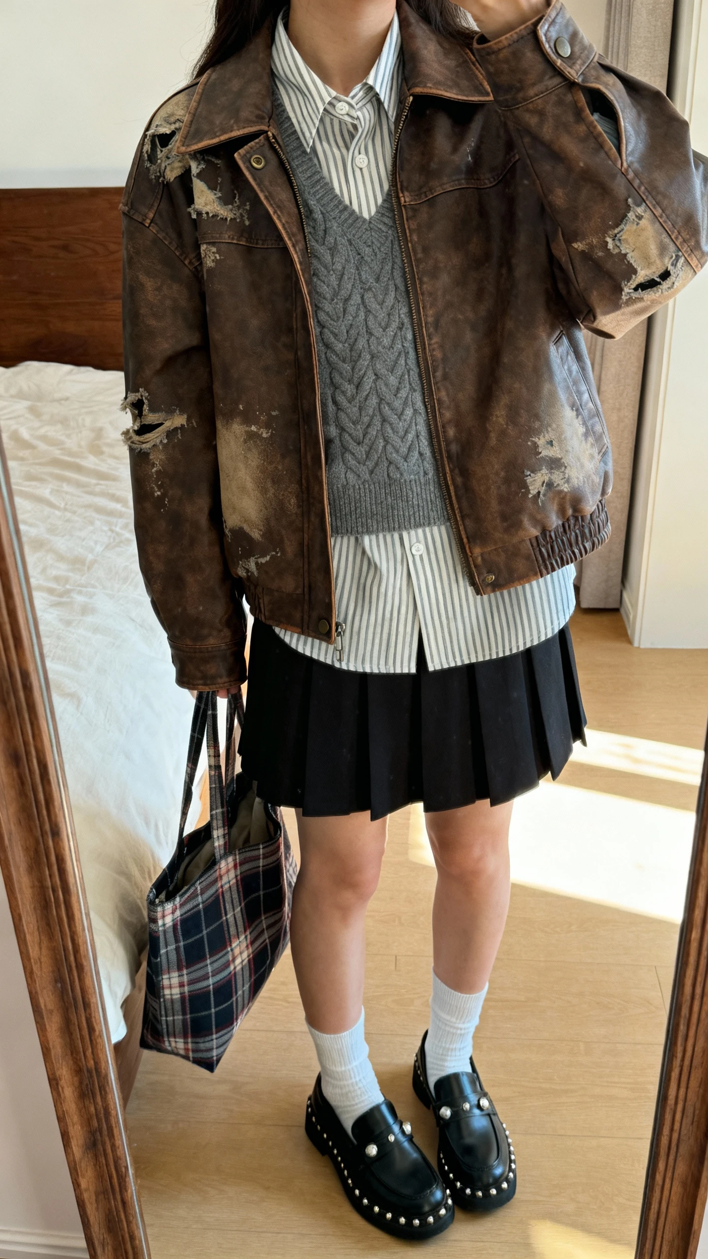 iPhone mirror selfie of a woman wearing a distressed brown leather jacket over a striped button-up shirt with a gray cable-knit vest layered on top, pleated black mini skirt, black loafers with white crew socks, silver studs, and a plaid tote, face not visible, cropped at neck, bedroom mirror, indirect afternoon light, iPhone photo quality
