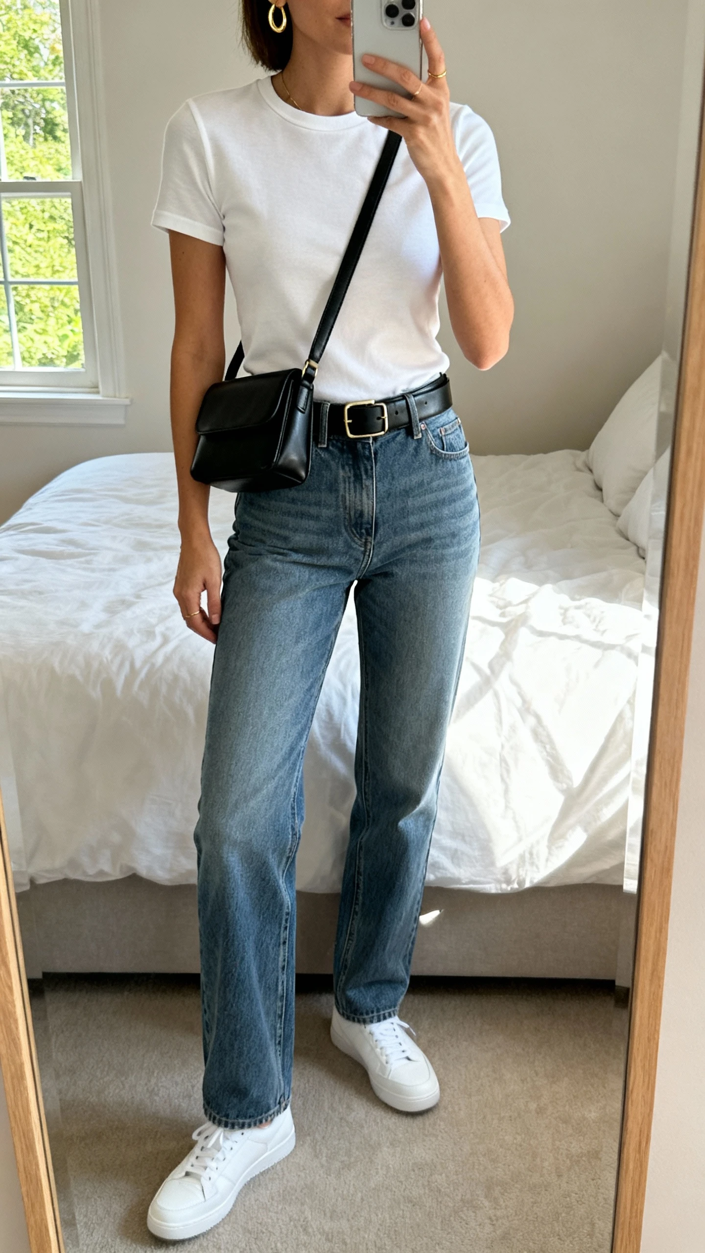 iPhone mirror selfie of a woman wearing a fitted white crewneck tee tucked into high-rise straight-leg medium-wash jeans, sleek black leather belt, minimalist white sneakers, delicate gold hoops, and a structured black crossbody bag, face not visible, cropped at neck, bedroom mirror with natural window light, iPhone photo quality.