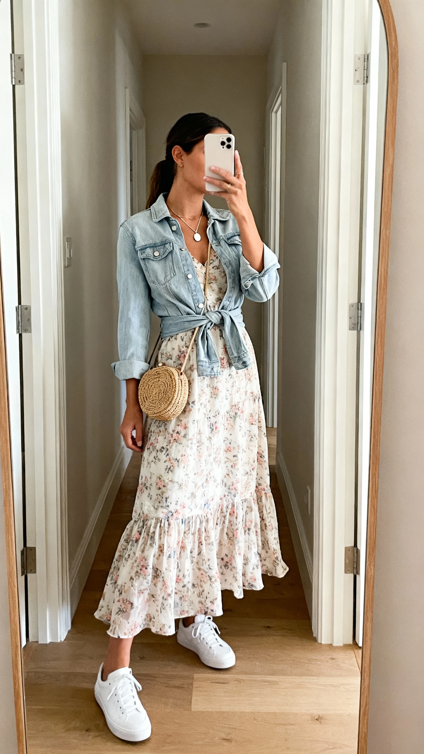 iPhone mirror selfie of a woman wearing a floaty floral midi dress paired with classic white sneakers, light denim jacket tied at the waist, delicate pendant necklace, and a woven mini bag, face not visible, side angle in a hallway full-length mirror with natural daylight, iPhone photo quality.