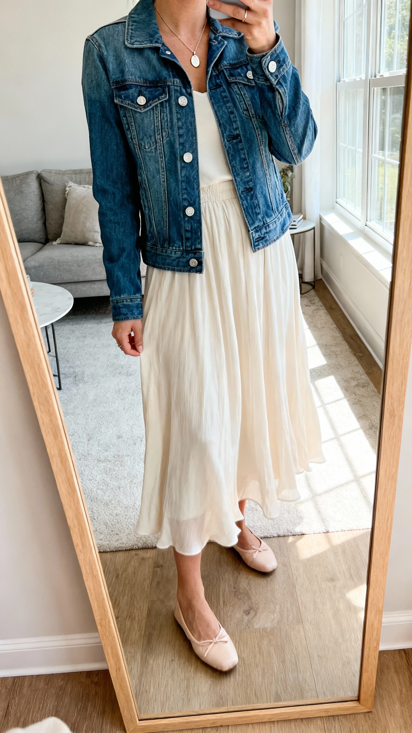 iPhone mirror selfie of a woman wearing a flowing midi skirt, classic denim jacket, and ballet flats with a delicate pendant necklace, face not visible, cropped at neck, living room full-length mirror with natural window light, iPhone photo quality.