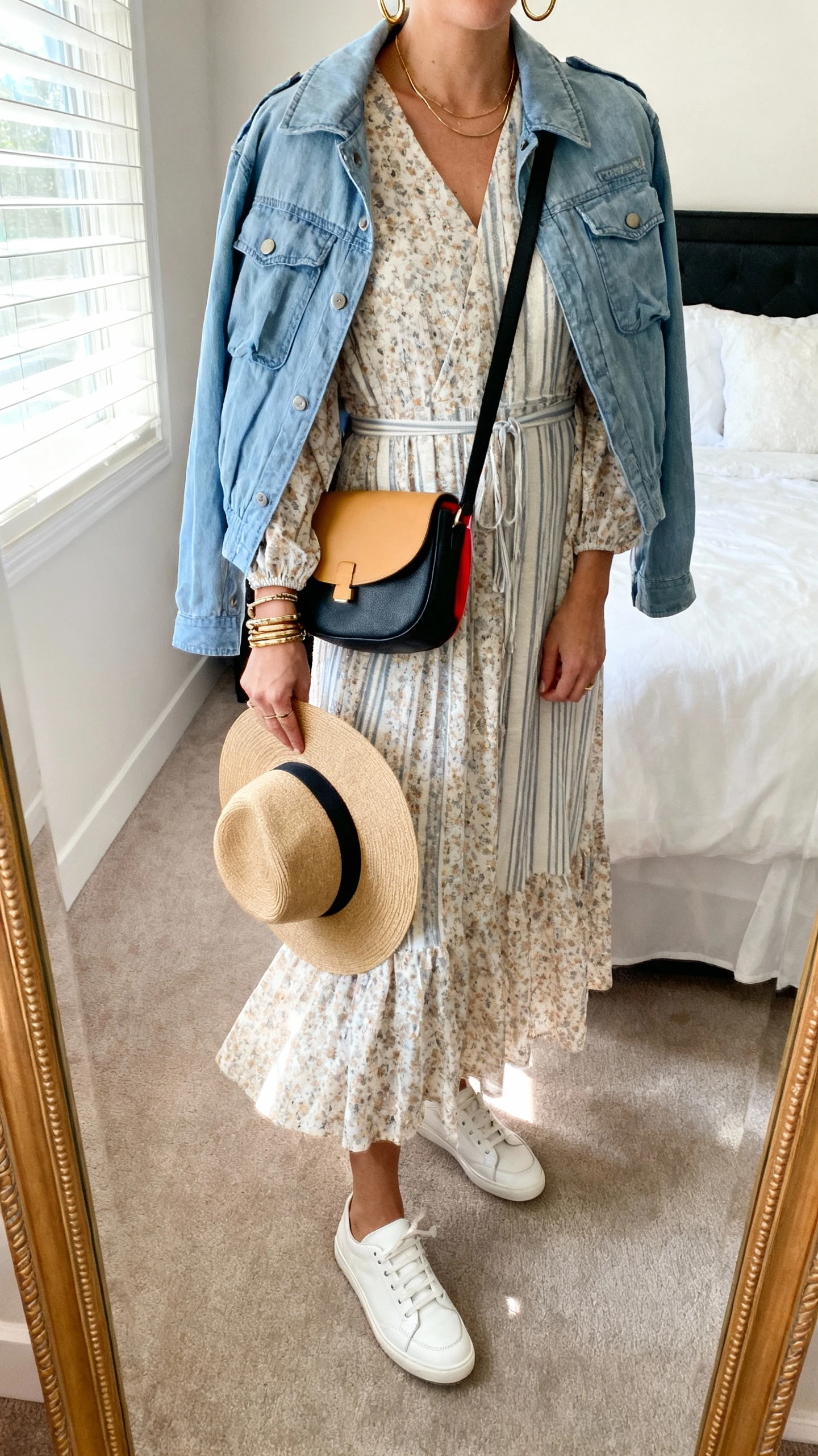 iPhone mirror selfie of a woman wearing a flowy midi dress (floral/stripe/solid), white leather sneakers with a clean silhouette, a denim or lightweight utility jacket, a tan/black/pop-color crossbody bag, minimal gold hoops, a stack of bracelets, and an optional wide-brim hat in hand, face not visible, cropped at neck, bedroom mirror with natural window light, iPhone photo quality.