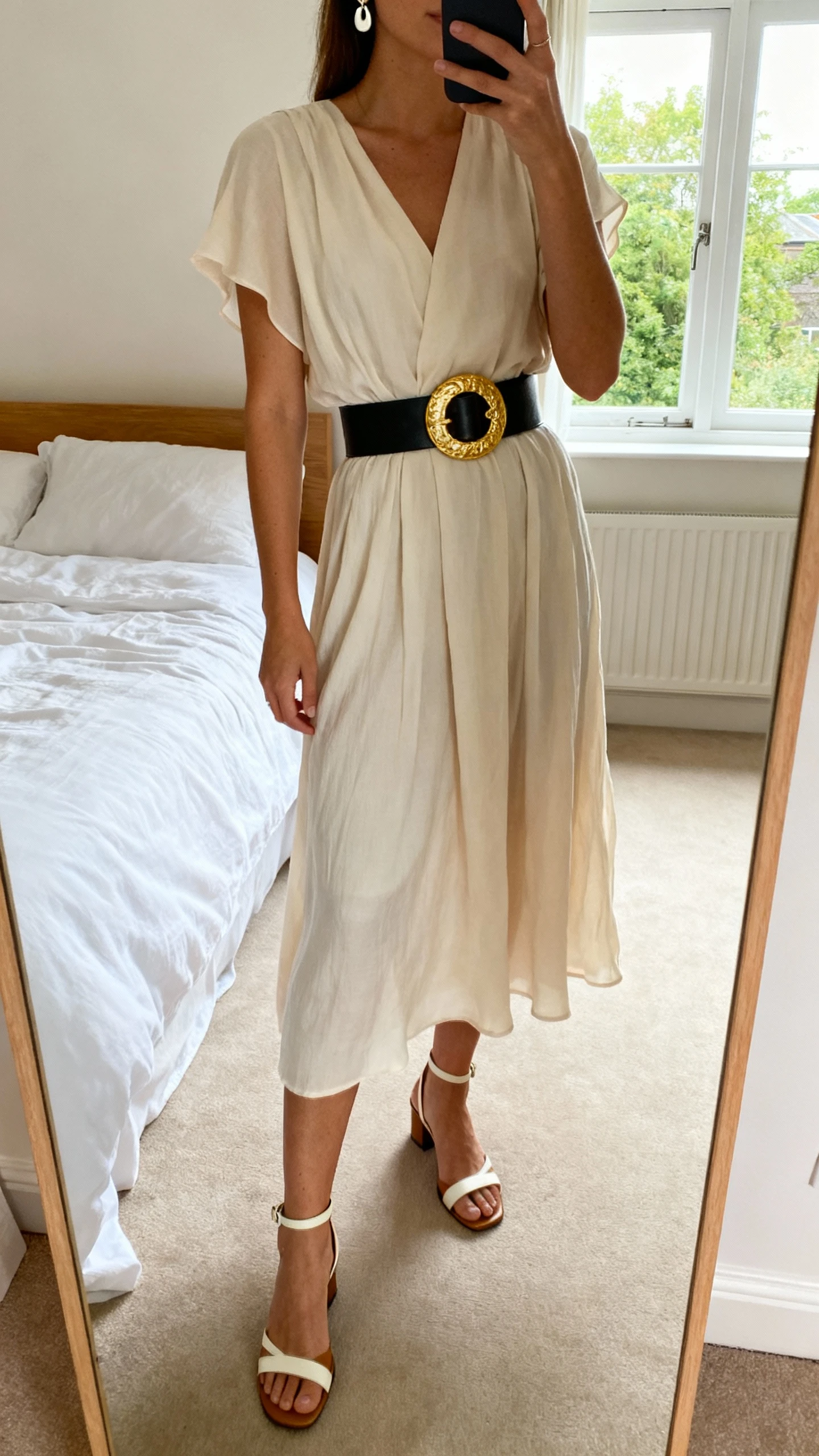 iPhone mirror selfie of a woman wearing a flowy midi dress cinched with a bold statement waist belt, simple ankle-strap heels, minimal earrings, face not visible, cropped at neck, bedroom mirror with natural window light, iPhone photo quality.
