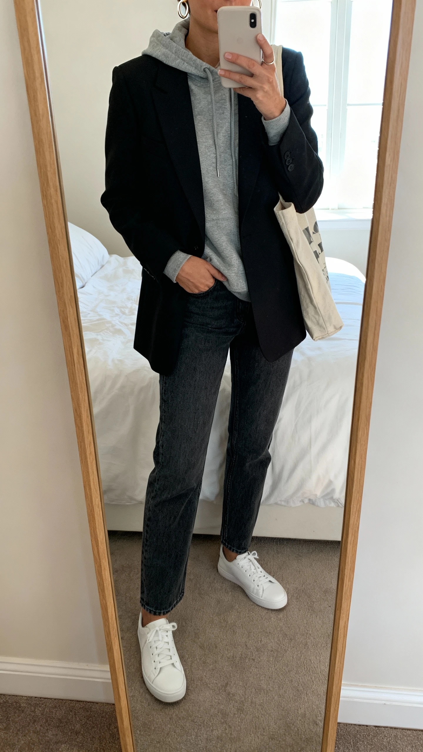 iPhone mirror selfie of a woman wearing a gray hoodie layered under a black tailored blazer, straight-leg denim, white low-top sneakers, simple hoop earrings, and a canvas tote, face not visible, cropped at neck, bedroom mirror, natural window light, iPhone photo quality.