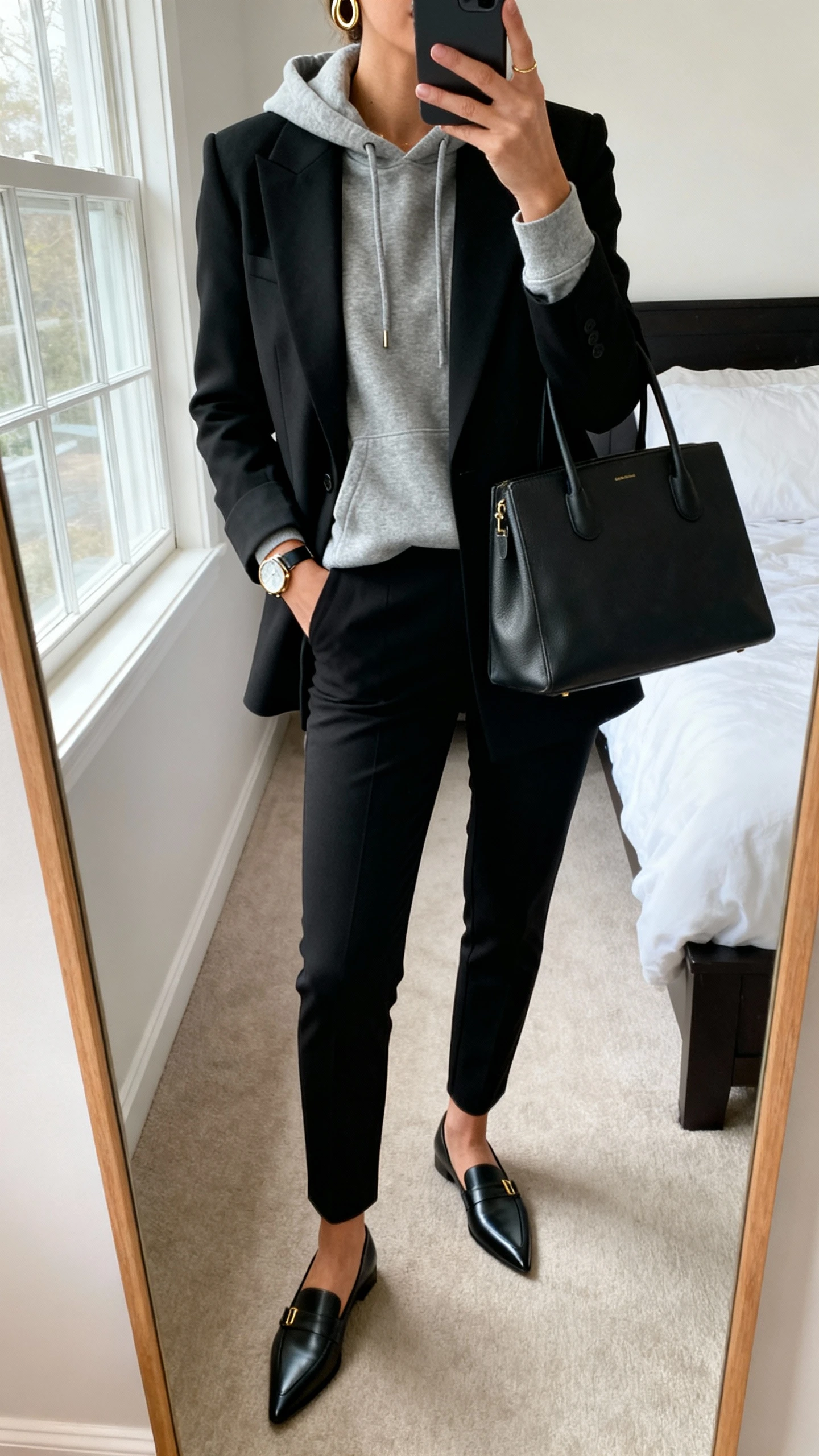 iPhone mirror selfie of a woman wearing a heather gray hoodie under a tailored black blazer, slim black trousers, pointed loafers, structured tote, minimal gold hoops and watch, face not visible, cropped at neck, bedroom mirror, natural window light, iPhone photo quality.