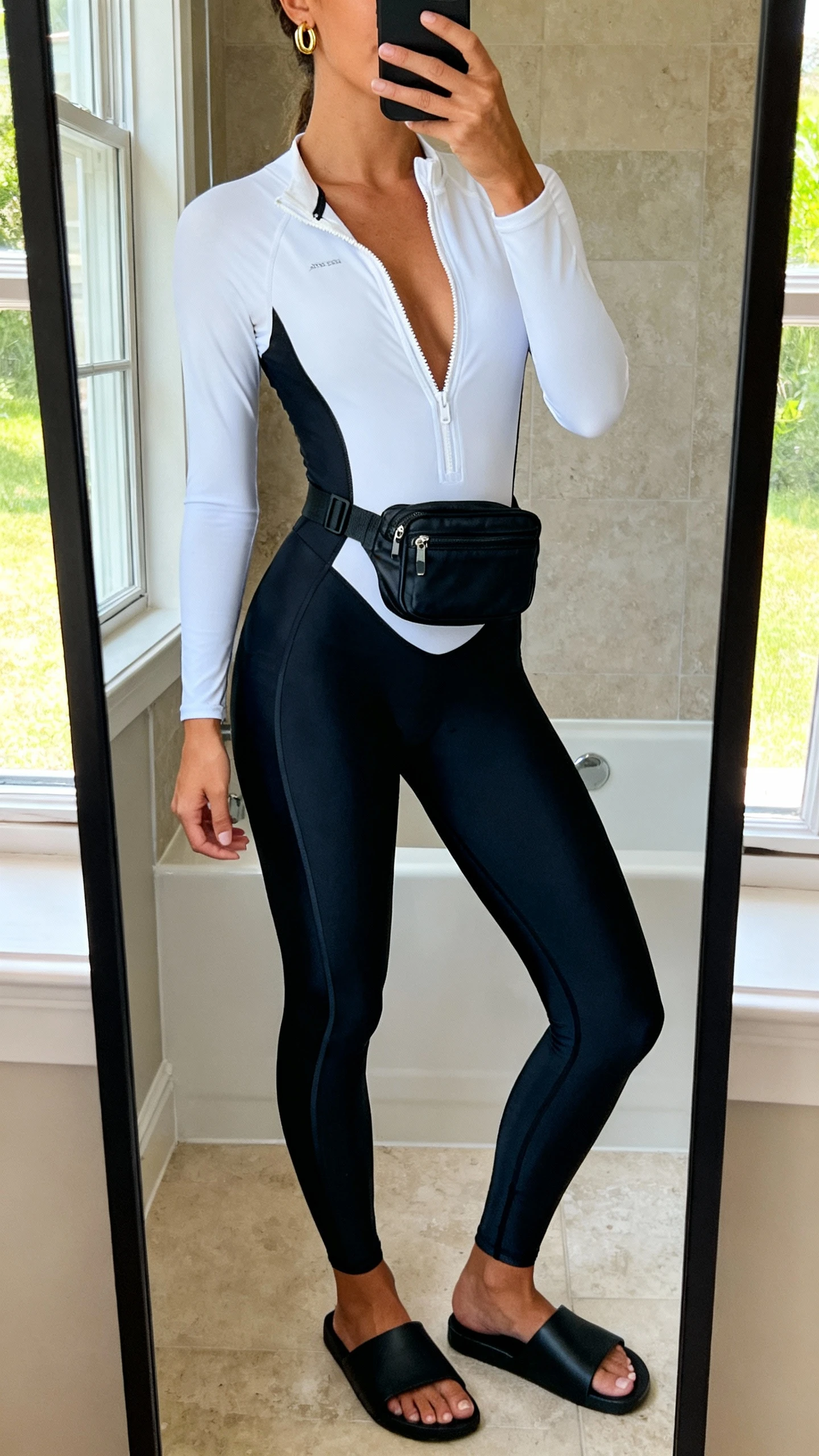 iPhone mirror selfie of a woman wearing a high-leg black monokini with sleek sporty lines, paired with a white zip-up rash guard half-open, minimal gold huggie earrings, black slide sandals, and a compact black belt bag, face not visible, cropped at neck, bathroom full-length mirror, morning window light, iPhone photo quality.