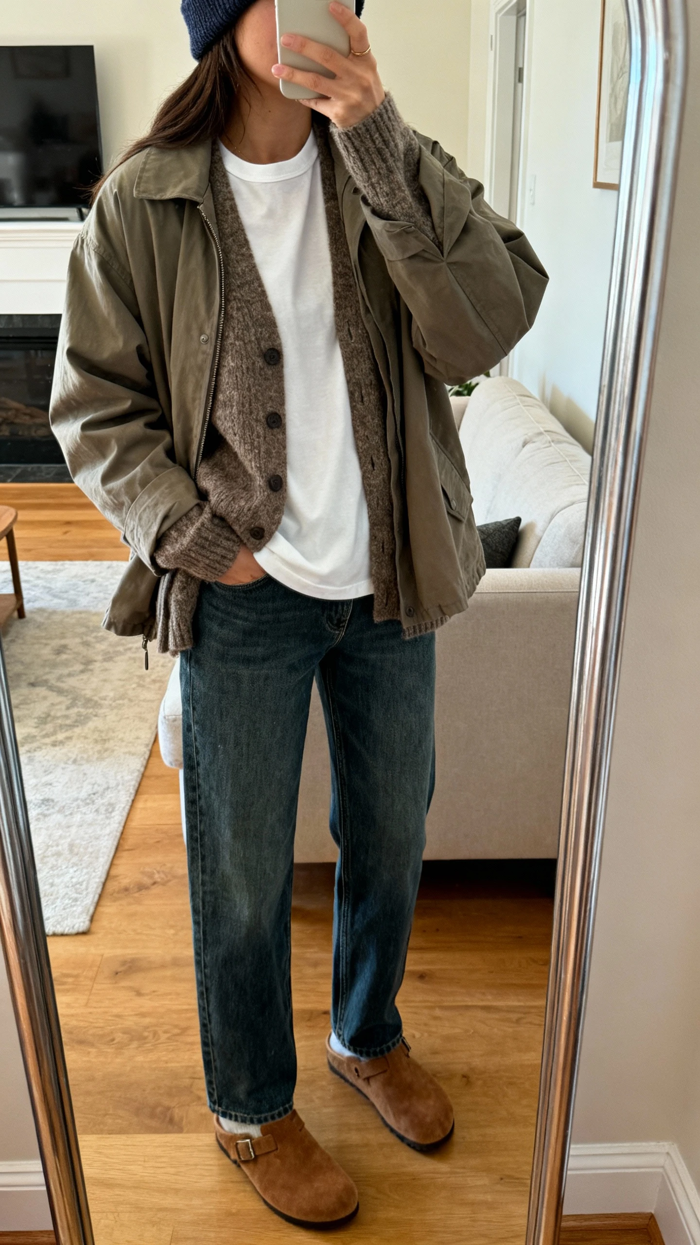 iPhone mirror selfie of a woman wearing a layered crew tee under a cozy cardigan topped with a relaxed shacket, straight jeans, suede clogs, and a beanie, face not visible, cropped at shoulders, living room full-length mirror, warm indoor lighting, iPhone photo quality.