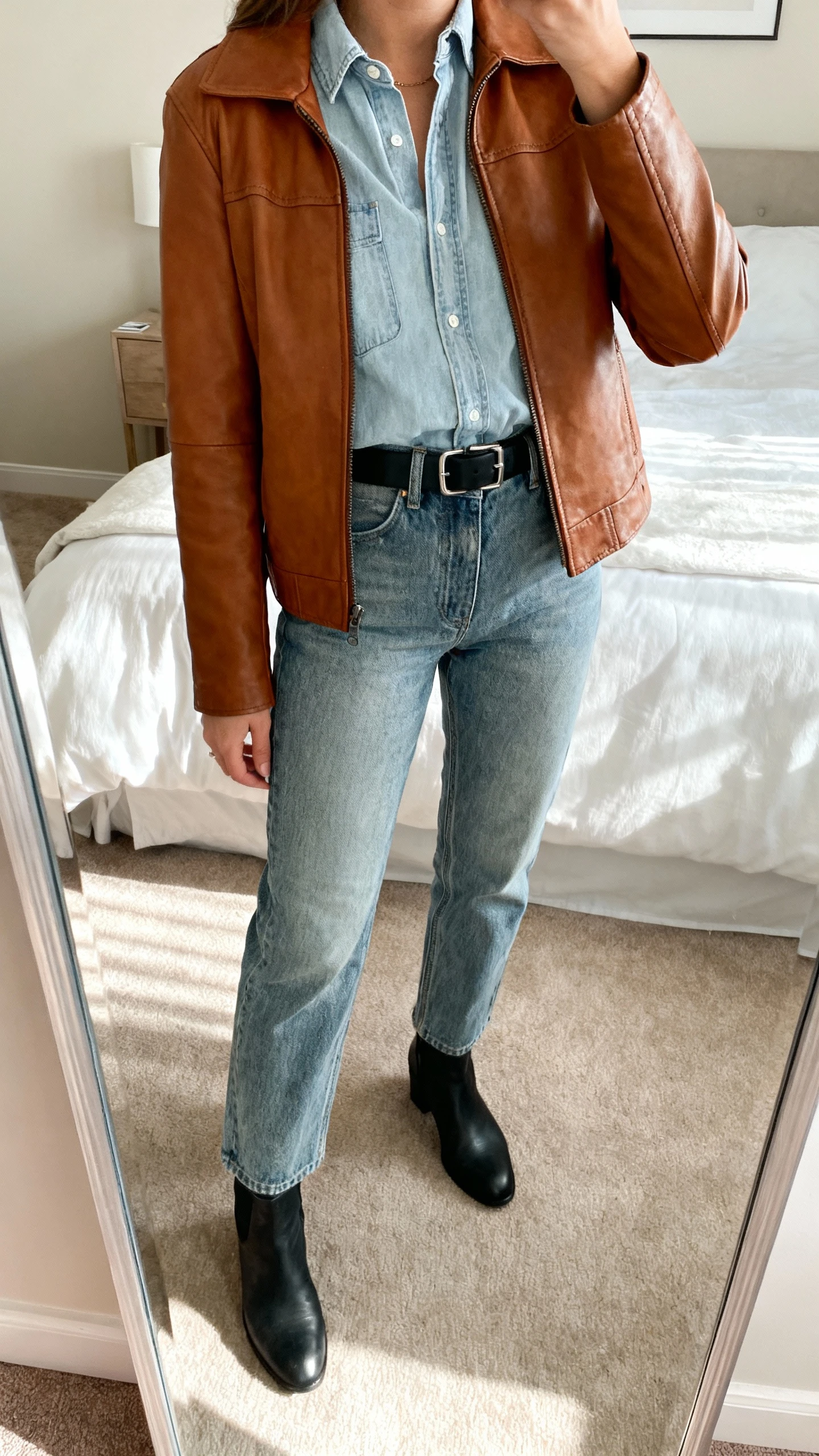 iPhone mirror selfie of a woman wearing a light denim shirt tucked into mid-wash straight denim jeans with a cognac brown leather jacket to ground the look, black belt, and simple black ankle boots, face not visible, cropped at shoulders, bedroom mirror, natural afternoon light, iPhone photo quality.