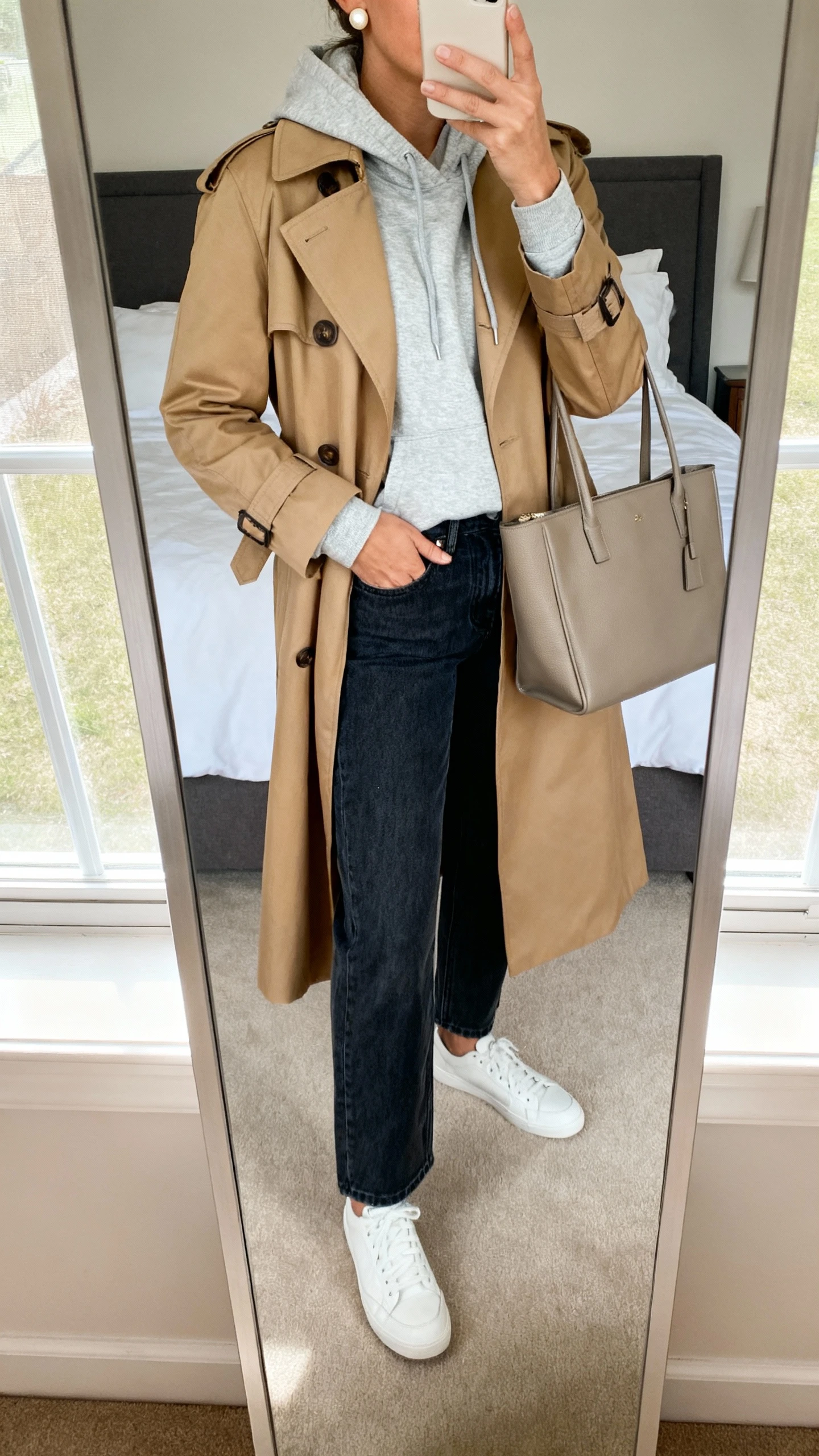 iPhone mirror selfie of a woman wearing a light gray hoodie under a classic tan trench coat, dark-wash straight-leg jeans, white sneakers, structured tote, simple pearl studs, face not visible, cropped at shoulders, bedroom mirror, natural window light, iPhone photo quality.