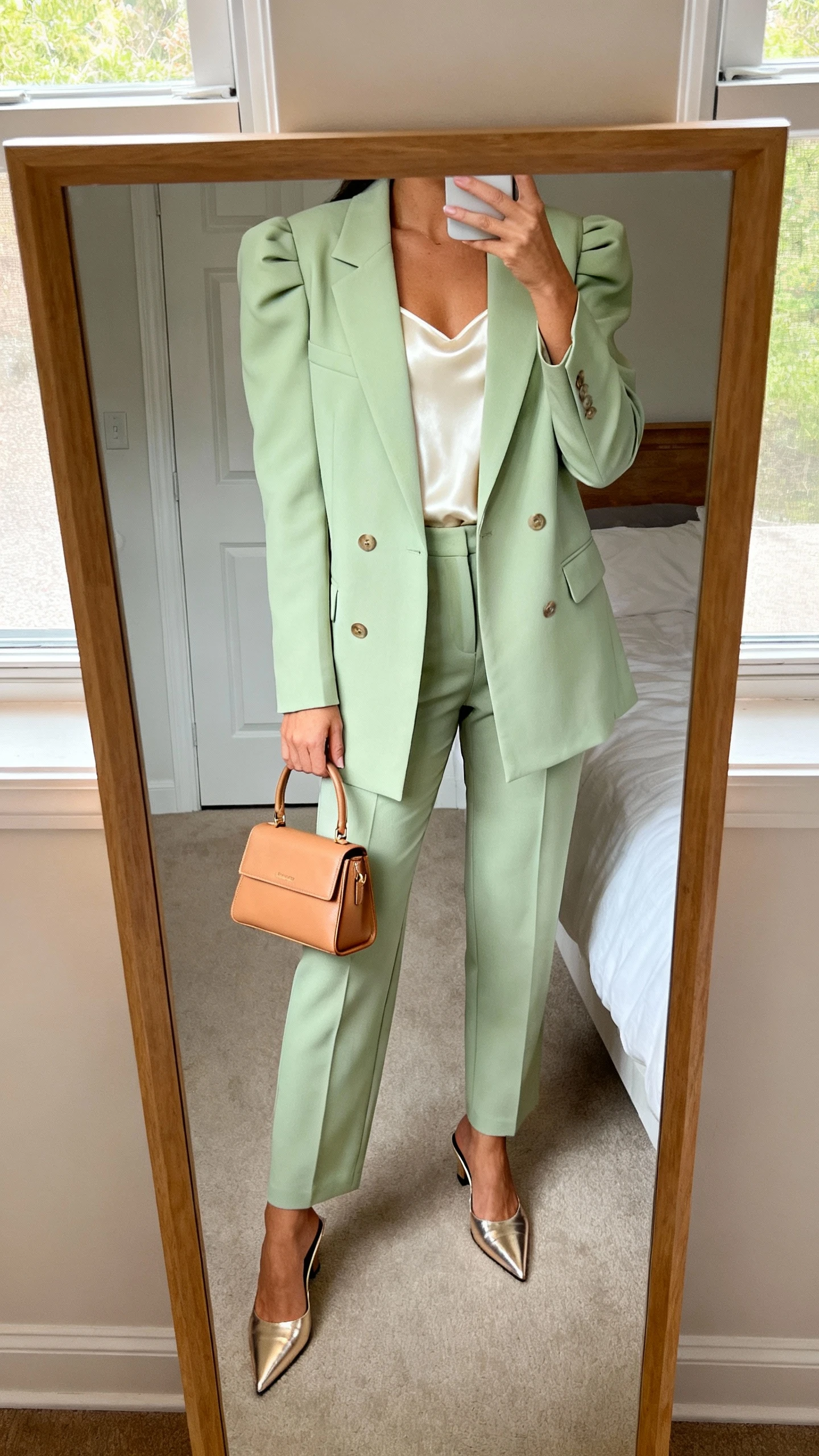 iPhone mirror selfie of a woman wearing a light pistachio single-breasted blazer with lightly padded shoulders, matching high-waisted tailored trousers, a silky cream camisole, pointed-toe mules in metallic champagne, and a structured mini tote in tan, face not visible, cropped at neck, bedroom mirror, natural window light, iPhone photo quality.
