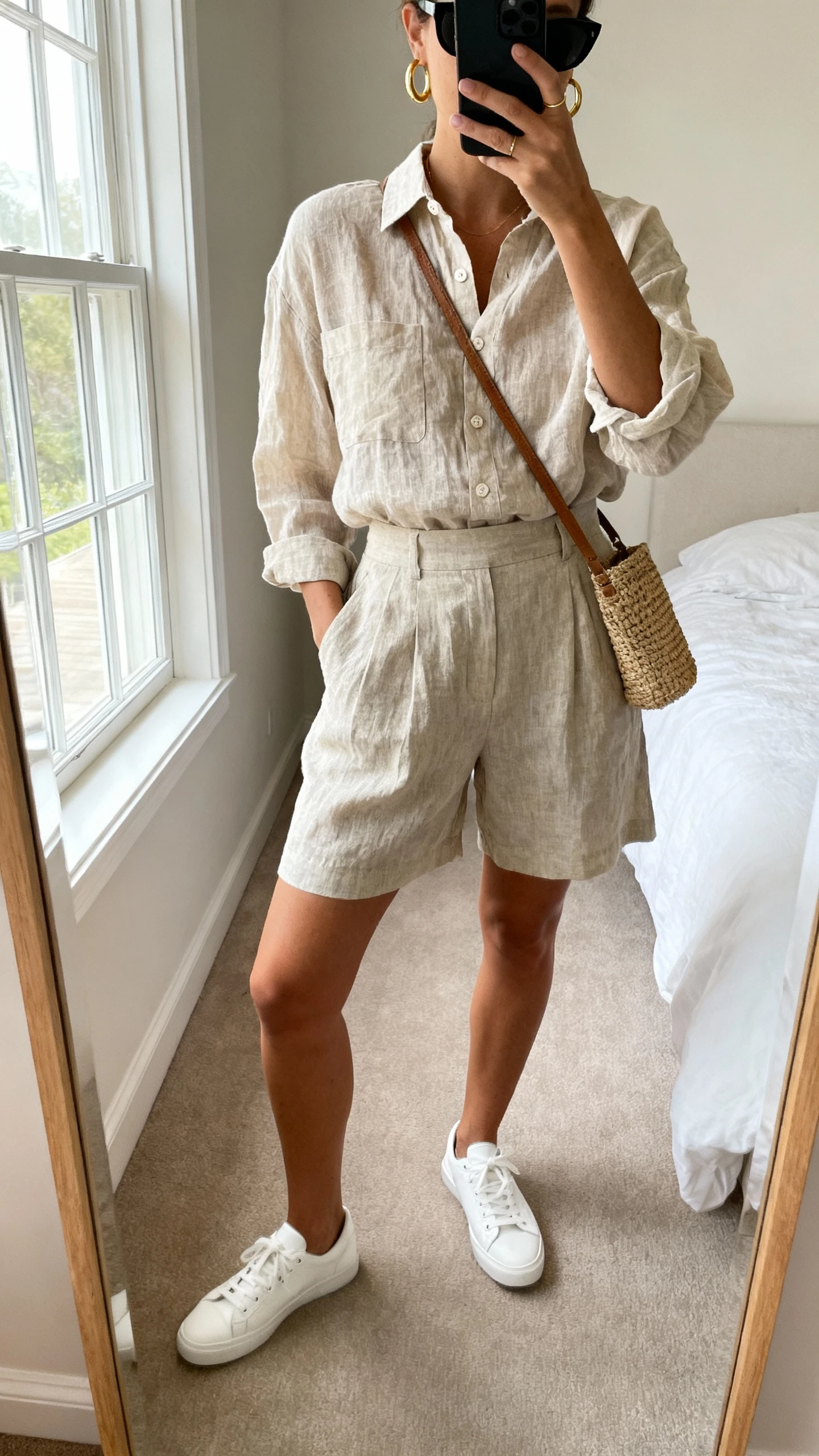 iPhone mirror selfie of a woman wearing a lightweight linen two-piece set (linen button-up shirt and matching high-waisted shorts) with sleek white low-top sneakers, simple gold hoops, a woven crossbody bag, and black sunglasses, face not visible, cropped at neck, bedroom mirror with natural window light, iPhone photo quality