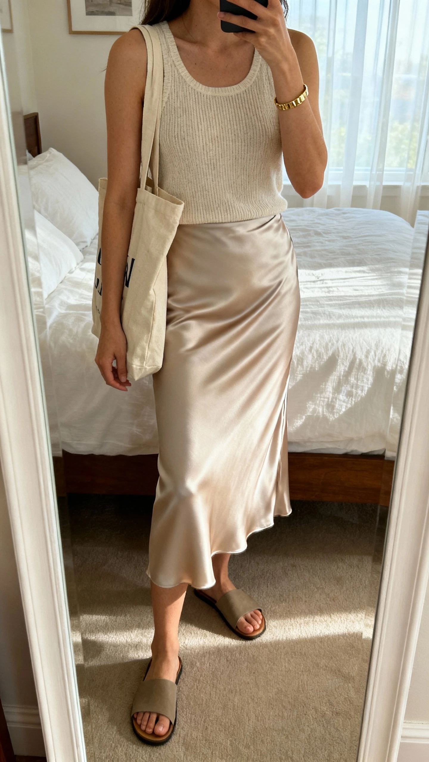iPhone mirror selfie of a woman wearing a lightweight knit tank and a satin bias-cut midi skirt, flat slide sandals, canvas tote, simple gold bracelet, face not visible, cropped at shoulders, bedroom mirror, soft afternoon window light, iPhone photo quality.
