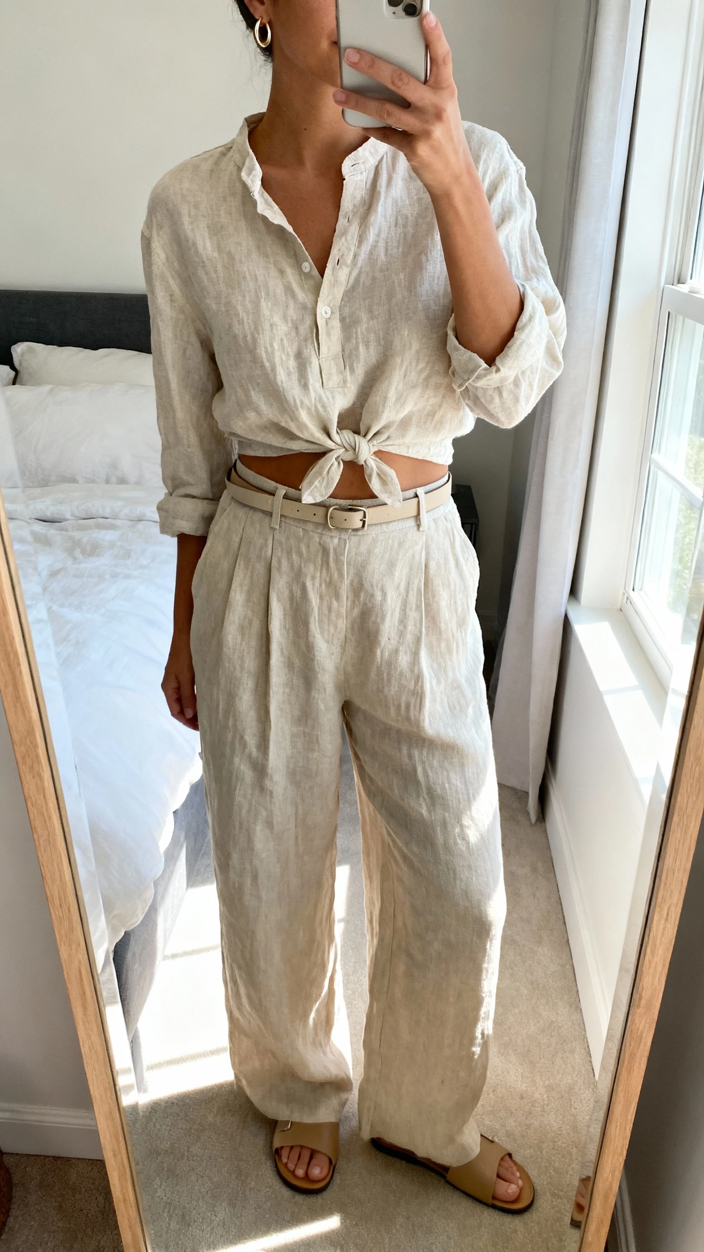 iPhone mirror selfie of a woman wearing a linen popover shirt half-tucked into high-waist wide-leg linen pants with minimalist leather slides, a slim belt, and small hoop earrings, face not visible, cropped at neck, bedroom mirror with bright window light, iPhone photo quality.