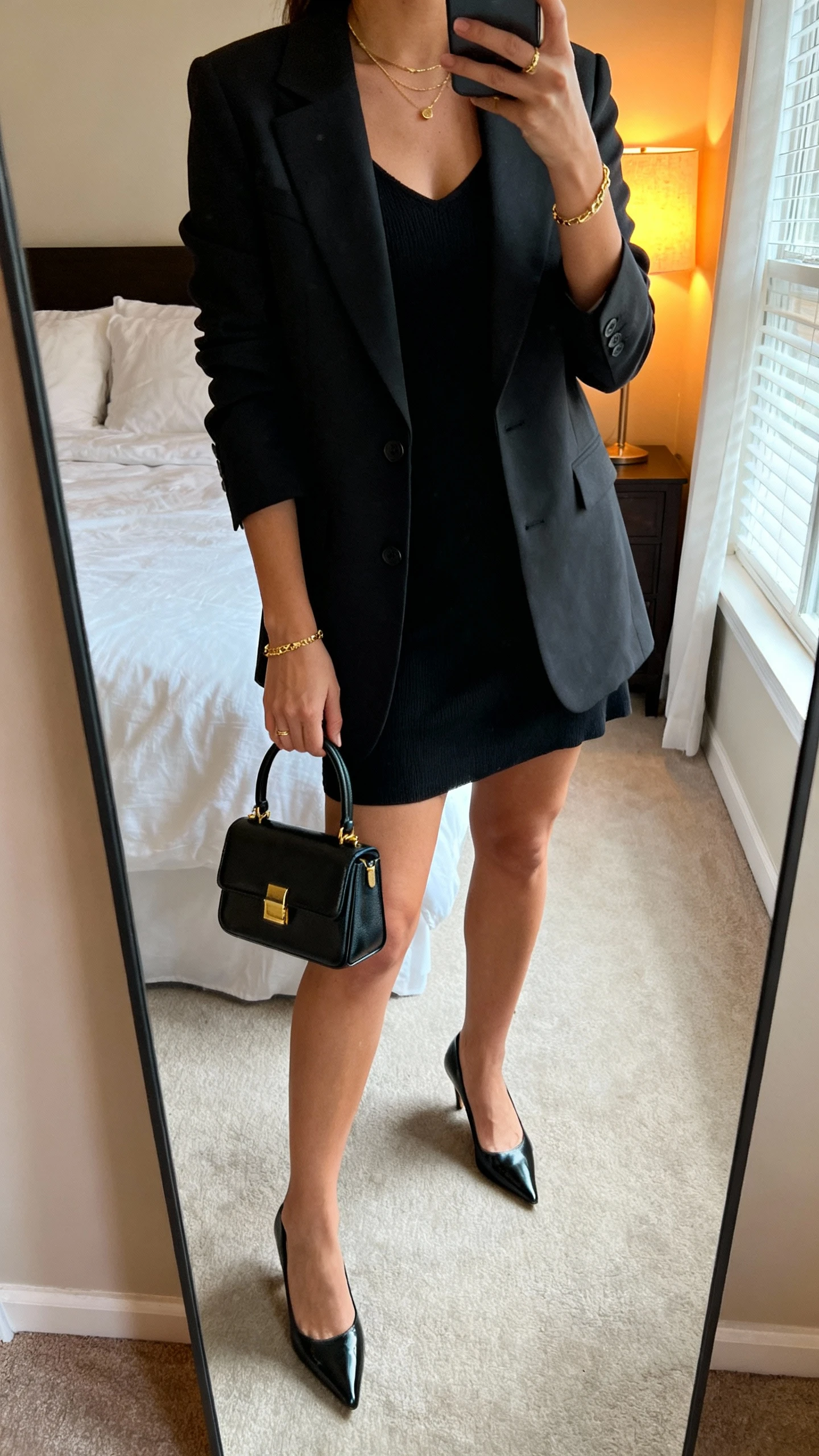 iPhone mirror selfie of a woman wearing a little black dress, tailored blazer, pointed heels, delicate gold jewelry, and carrying a structured mini top-handle bag, face not visible, cropped at neck, bedroom mirror with warm lamp light and window glow, iPhone photo quality.