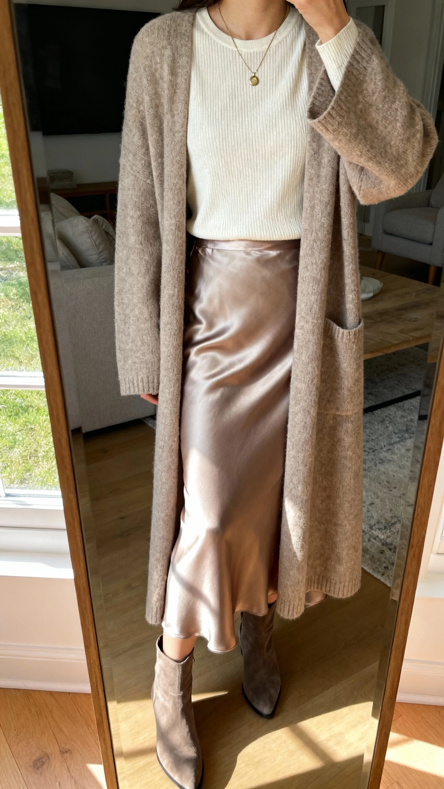 iPhone mirror selfie of a woman wearing a long cardigan coat over a satin slip skirt with a fitted knit tee and ankle boots, delicate pendant necklace, face not visible, cropped at shoulders, living-room mirror, soft afternoon light, iPhone photo quality.