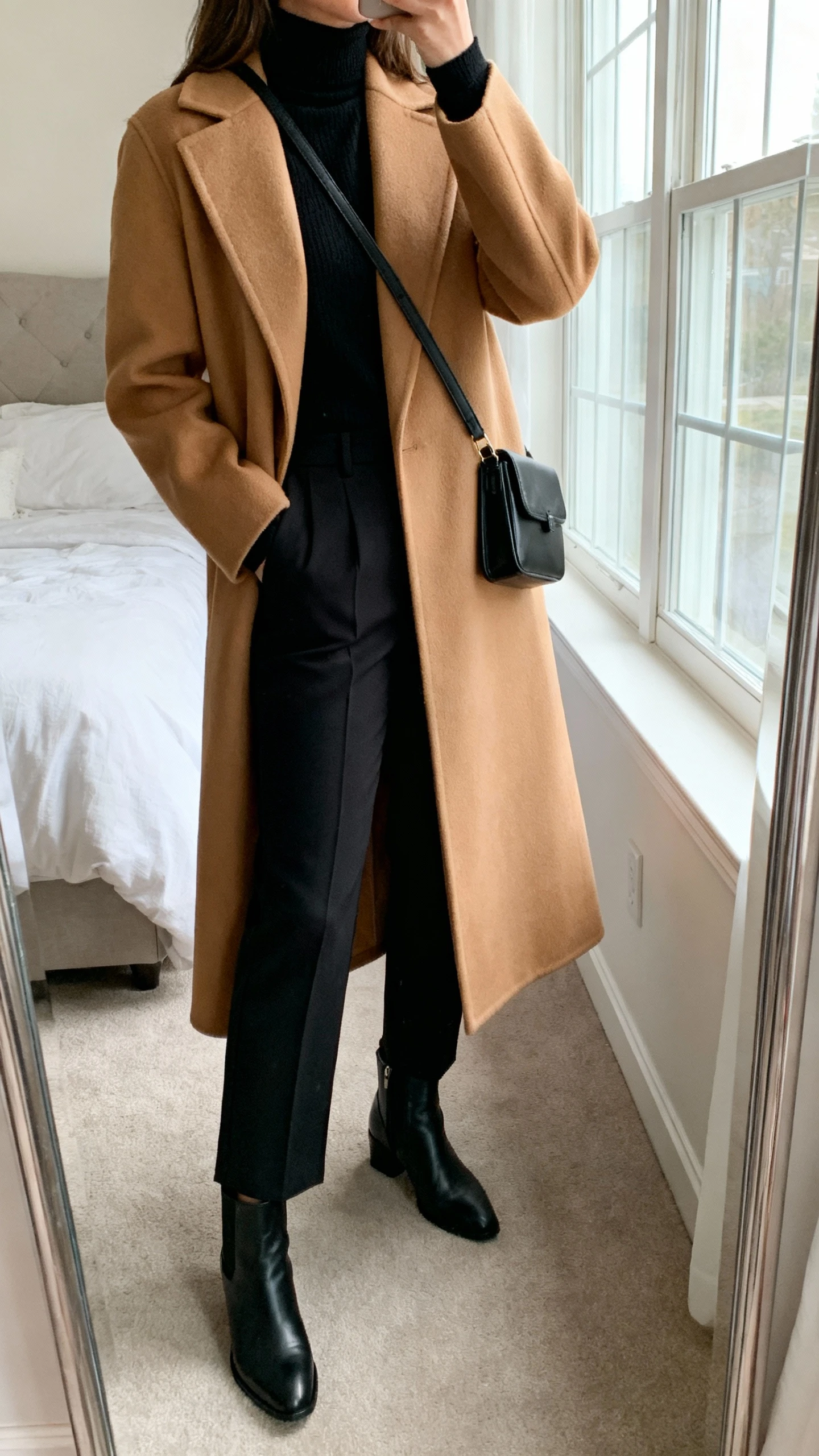 iPhone mirror selfie of a woman wearing a long structured camel power coat over an all-black monochrome base (black turtleneck, black tailored trousers), with sleek black ankle boots and a small black crossbody, face not visible, cropped at neck, bedroom mirror, natural window light, iPhone photo quality.