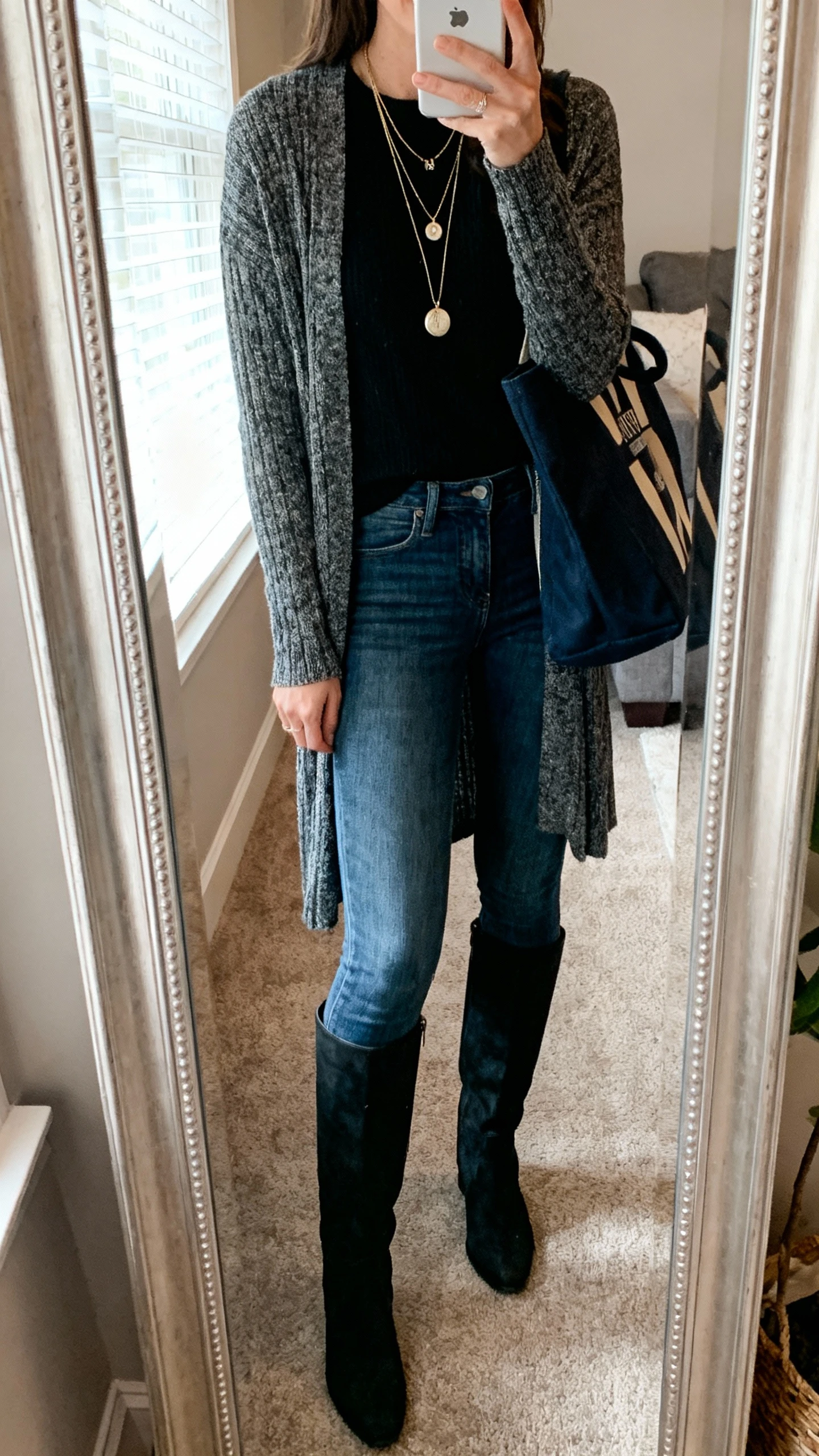 iPhone mirror selfie of a woman wearing a longline cardigan, skinny jeans that actually fit, and over-the-knee boots, face not visible, cropped at shoulders, living room mirror with window light, iPhone photo quality; layered pendant necklace and tote bag.