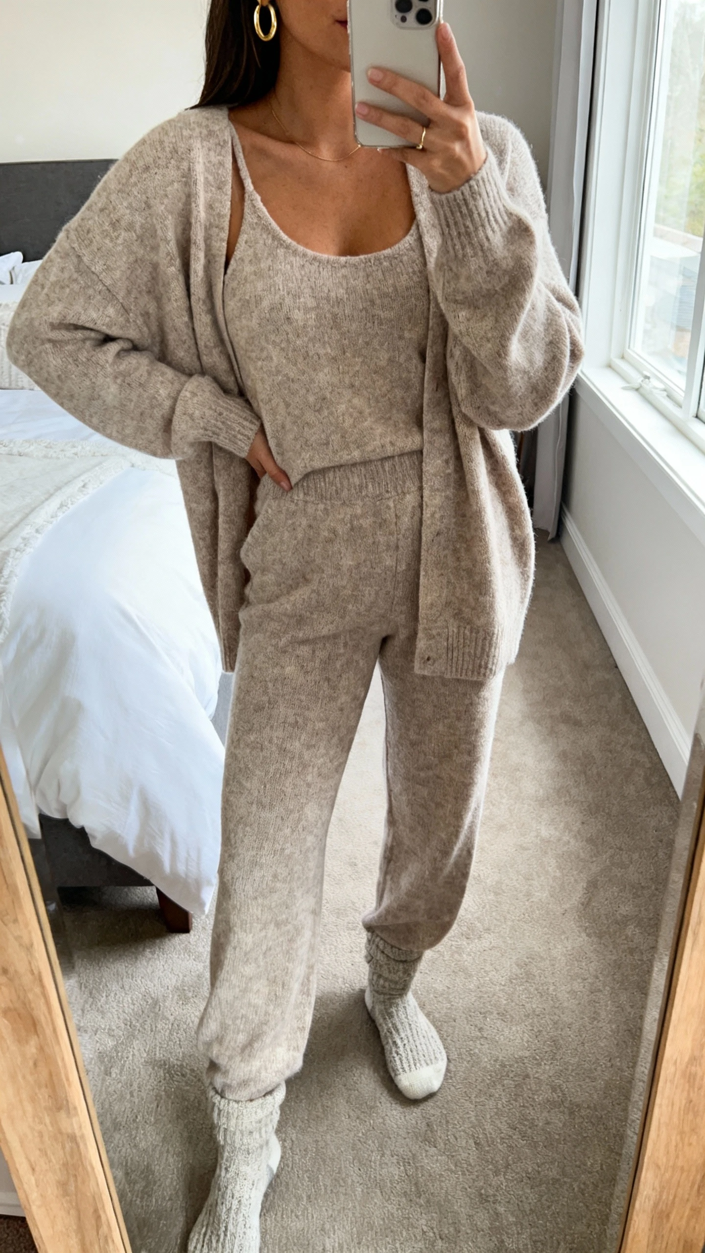iPhone mirror selfie of a woman wearing a luxe matching sweater set (soft knit cardigan + coordinating knit tank + matching knit pants) with minimalist gold hoops and cozy socks, face not visible, cropped at neck, bedroom mirror, natural window light, iPhone photo quality.