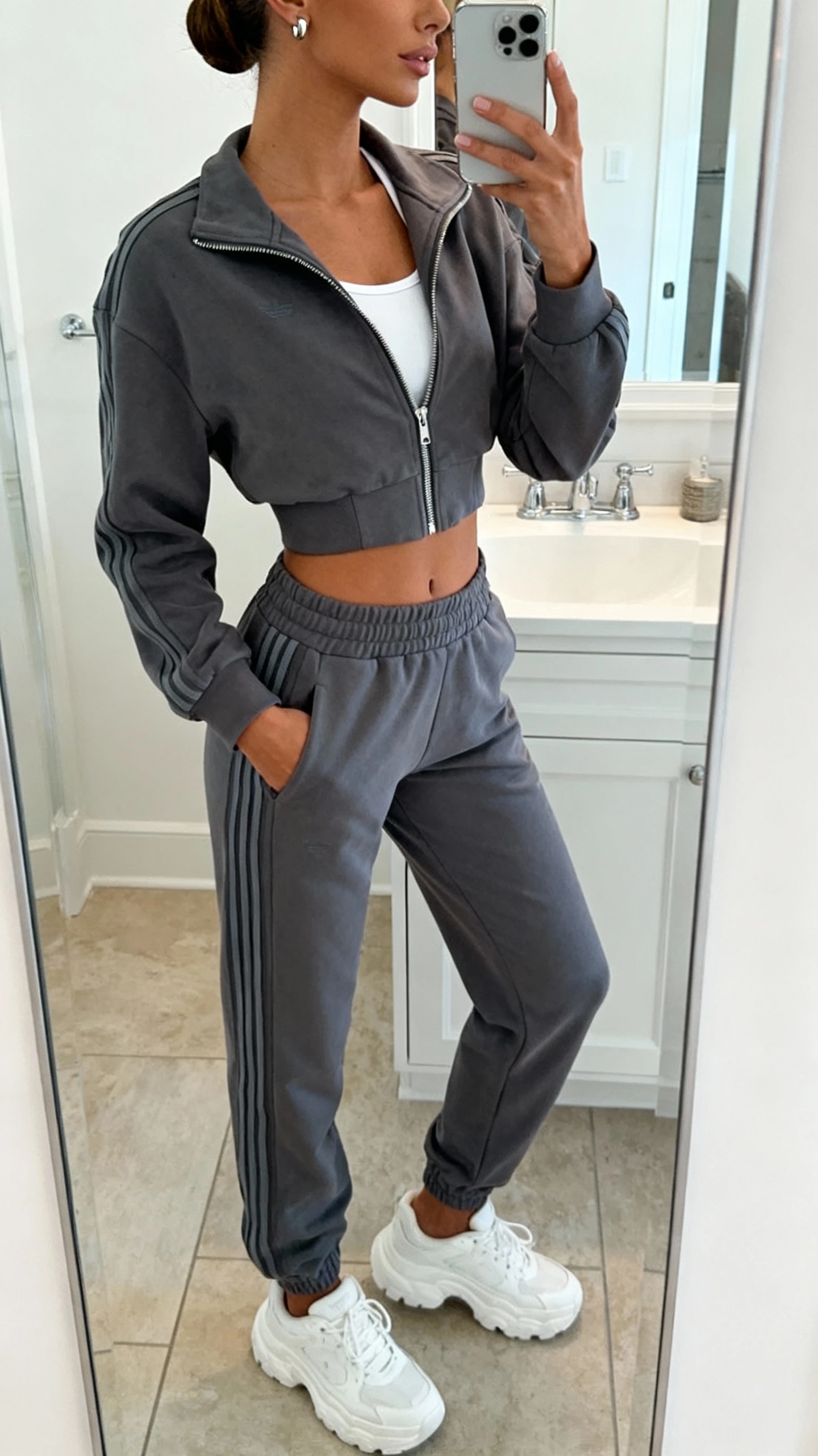 iPhone mirror selfie of a woman wearing a matching sporty luxe set: charcoal zip-up track jacket and high-waist track pants with subtle stripes, white crop tank underneath, chunky white trainers, sleek bun, and silver stud earrings; face not visible, cropped at neck, bathroom mirror, bright indoor lighting, iPhone photo quality.