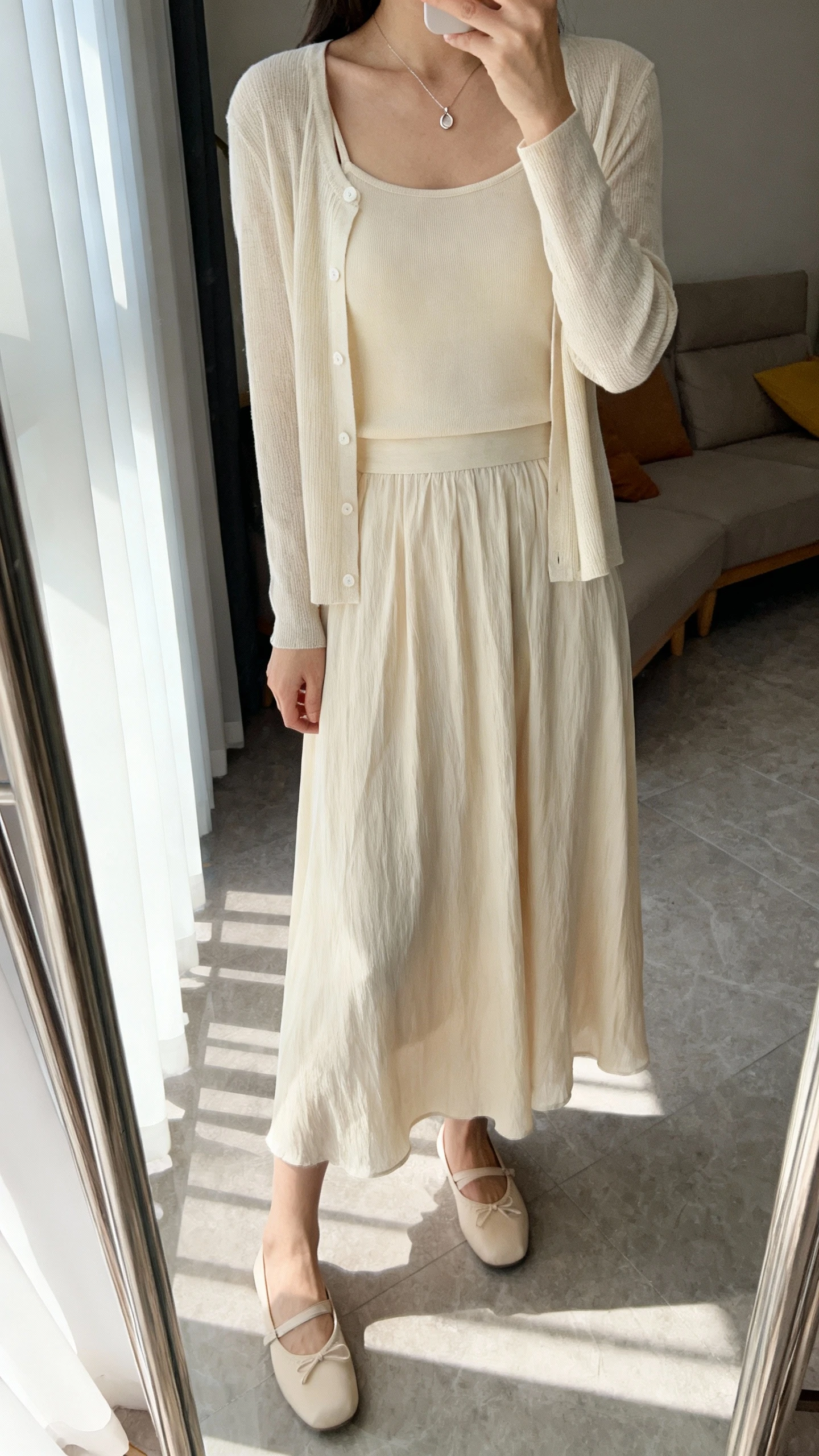 iPhone mirror selfie of a woman wearing a matching cardigan set (camisole + cardigan) with a flowy midi skirt, ballet flats, and a delicate pendant necklace, face not visible, cropped at neck, living room full-length mirror with window light, iPhone photo quality.