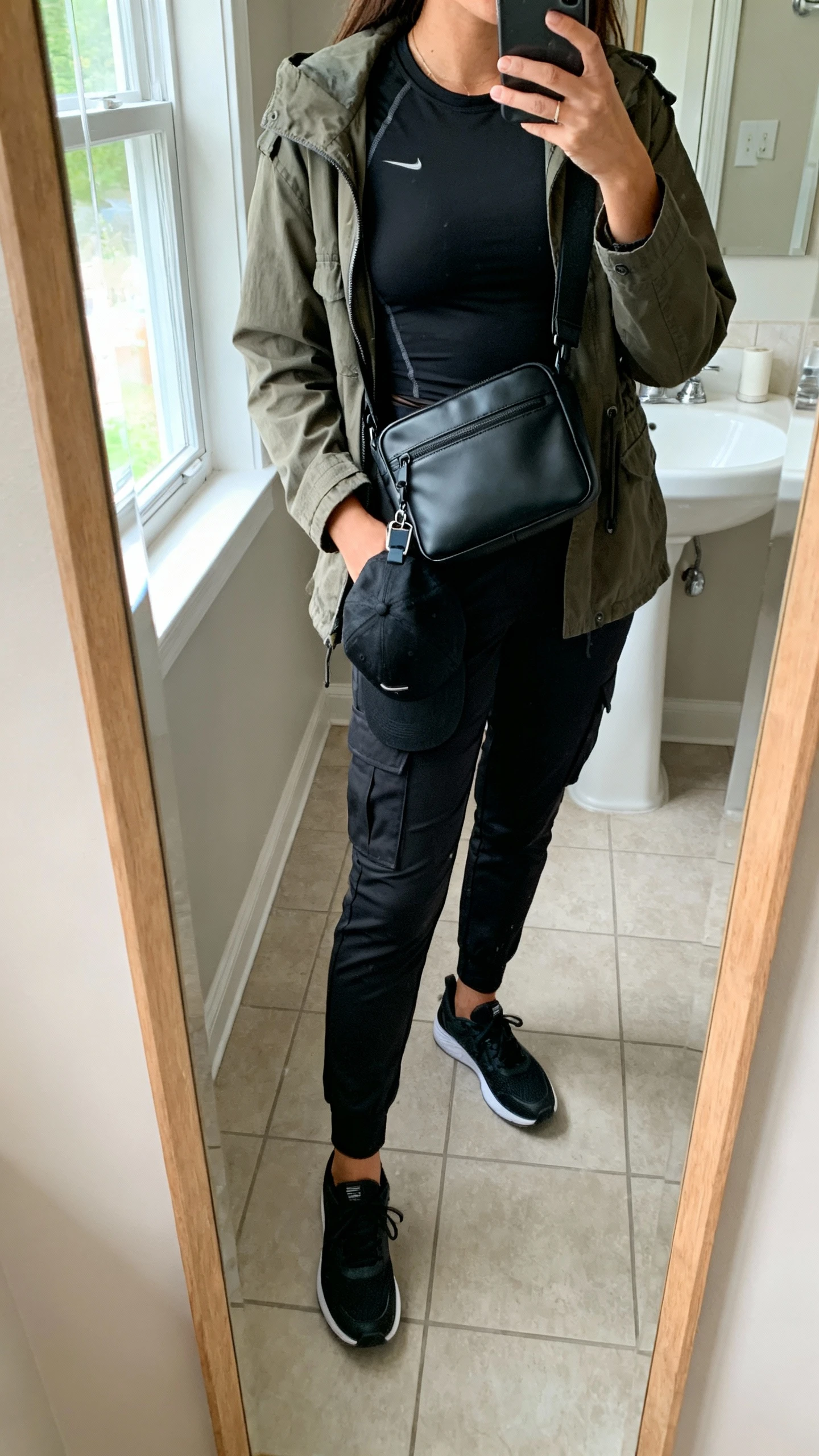 iPhone mirror selfie of a woman wearing a matte black techy crossbody bag, utility jacket layered over a fitted black tech tee, tapered cargo pants, sleek black sneakers, and a black cap clipped to the bag; face not visible, cropped at neck, bathroom full-length mirror, morning window light, iPhone photo quality.