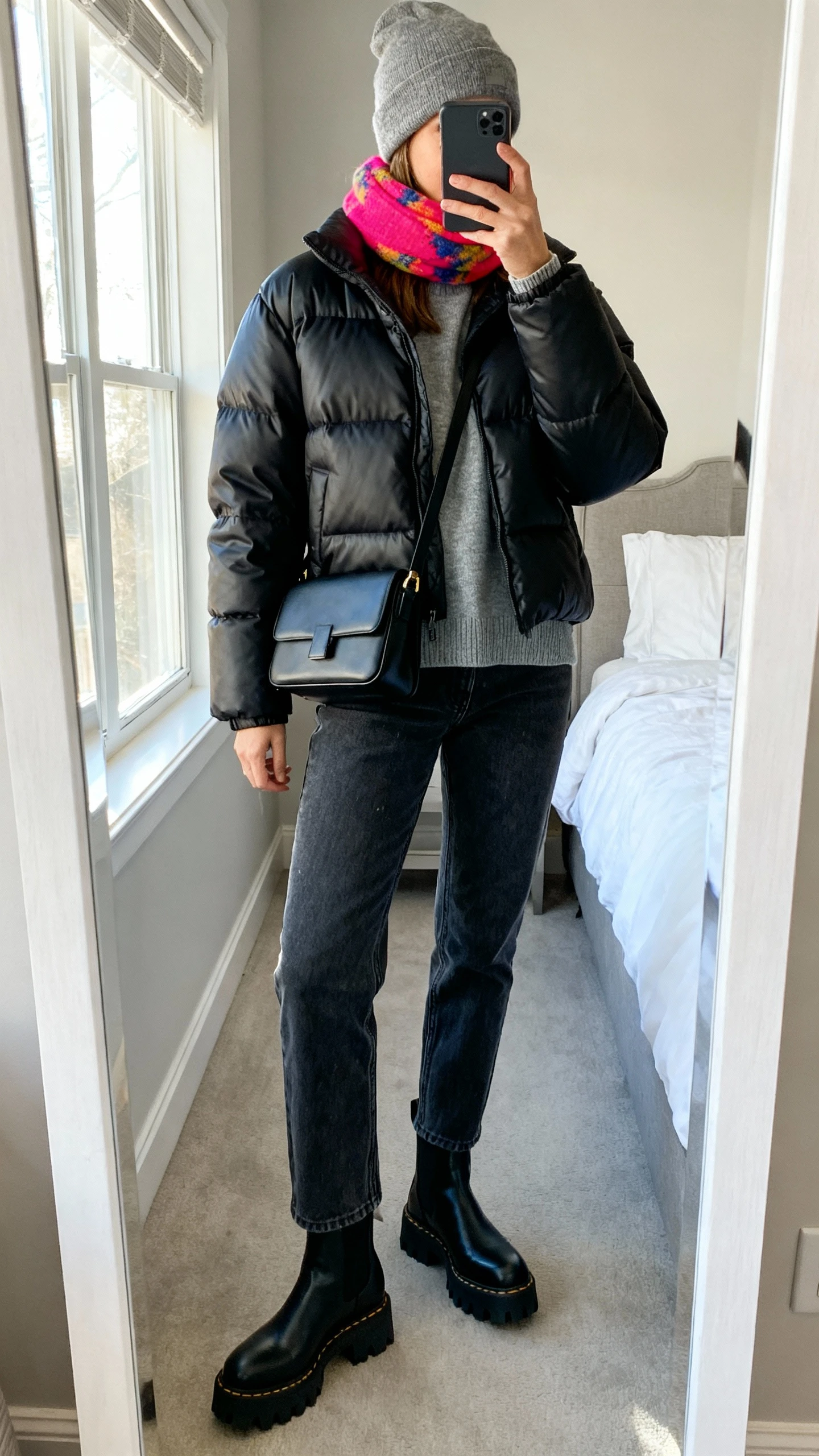 iPhone mirror selfie of a woman wearing a matte black puffer jacket (hip length), straight-leg dark-wash jeans, chunky black lug-sole ankle boots, a gray crewneck sweater, a structured black crossbody bag, and a heather gray beanie with a bright colorful scarf, face not visible, cropped at neck, bedroom mirror, natural window light, iPhone photo quality.