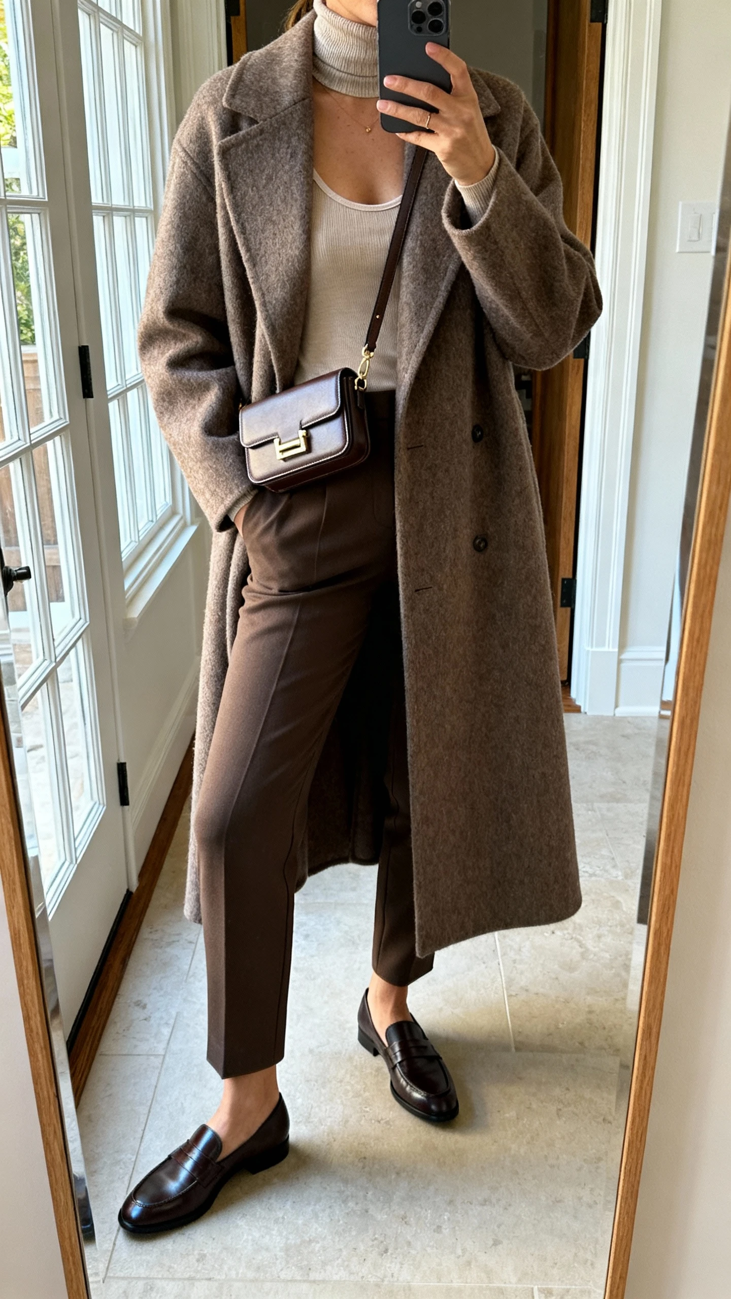 iPhone mirror selfie of a woman wearing a maxi wool overcoat over a thin turtleneck and a micro-layered camisole, with slim trousers, sleek loafers, and a compact crossbody, face not visible, cropped at neck, entryway full-length mirror, natural window light, iPhone photo quality.