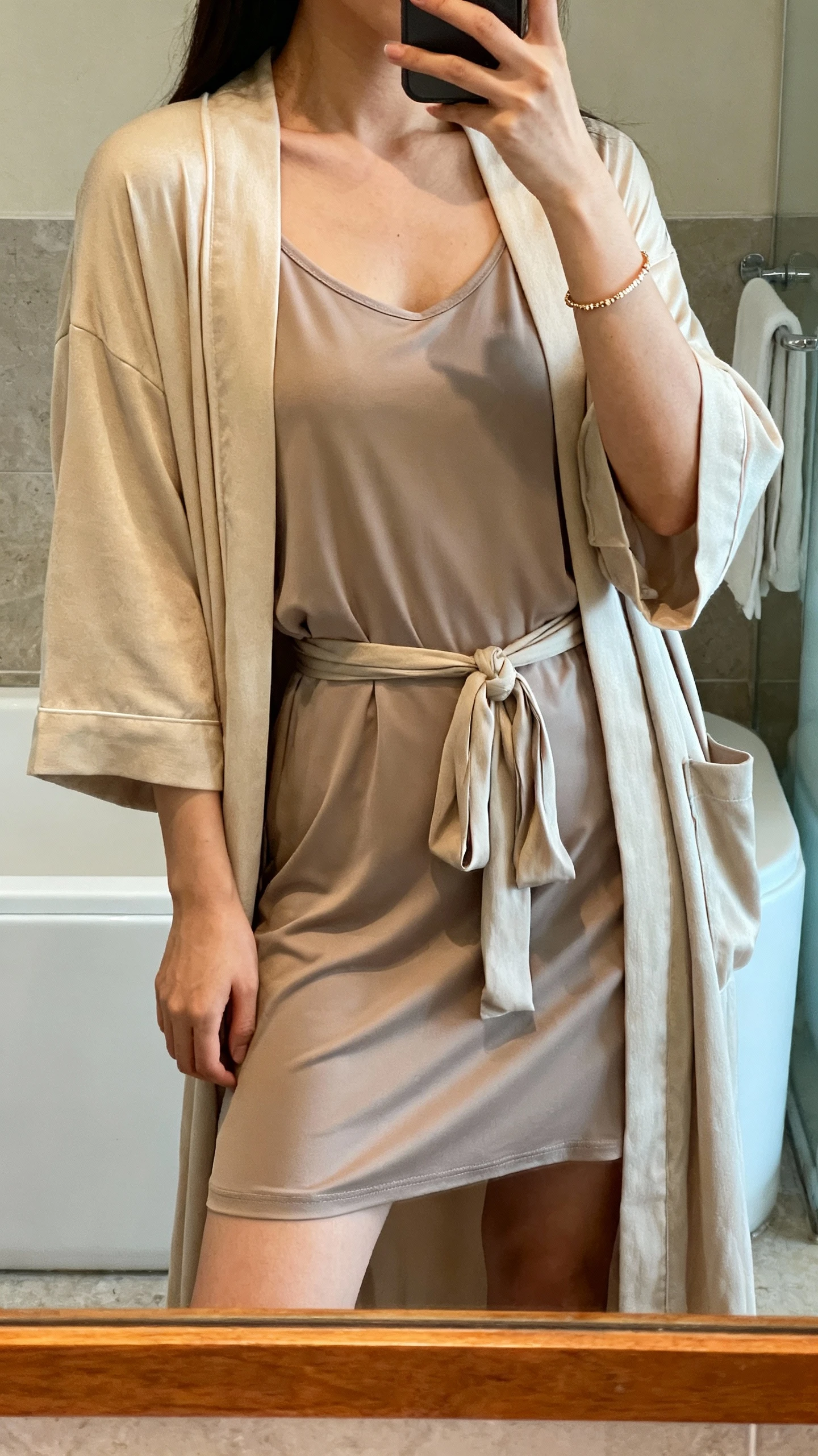 iPhone mirror selfie of a woman wearing a modal lounge dress with a matching robe tied loosely, delicate bracelet, face not visible, bathroom mirror, warm indoor lighting, iPhone photo quality.