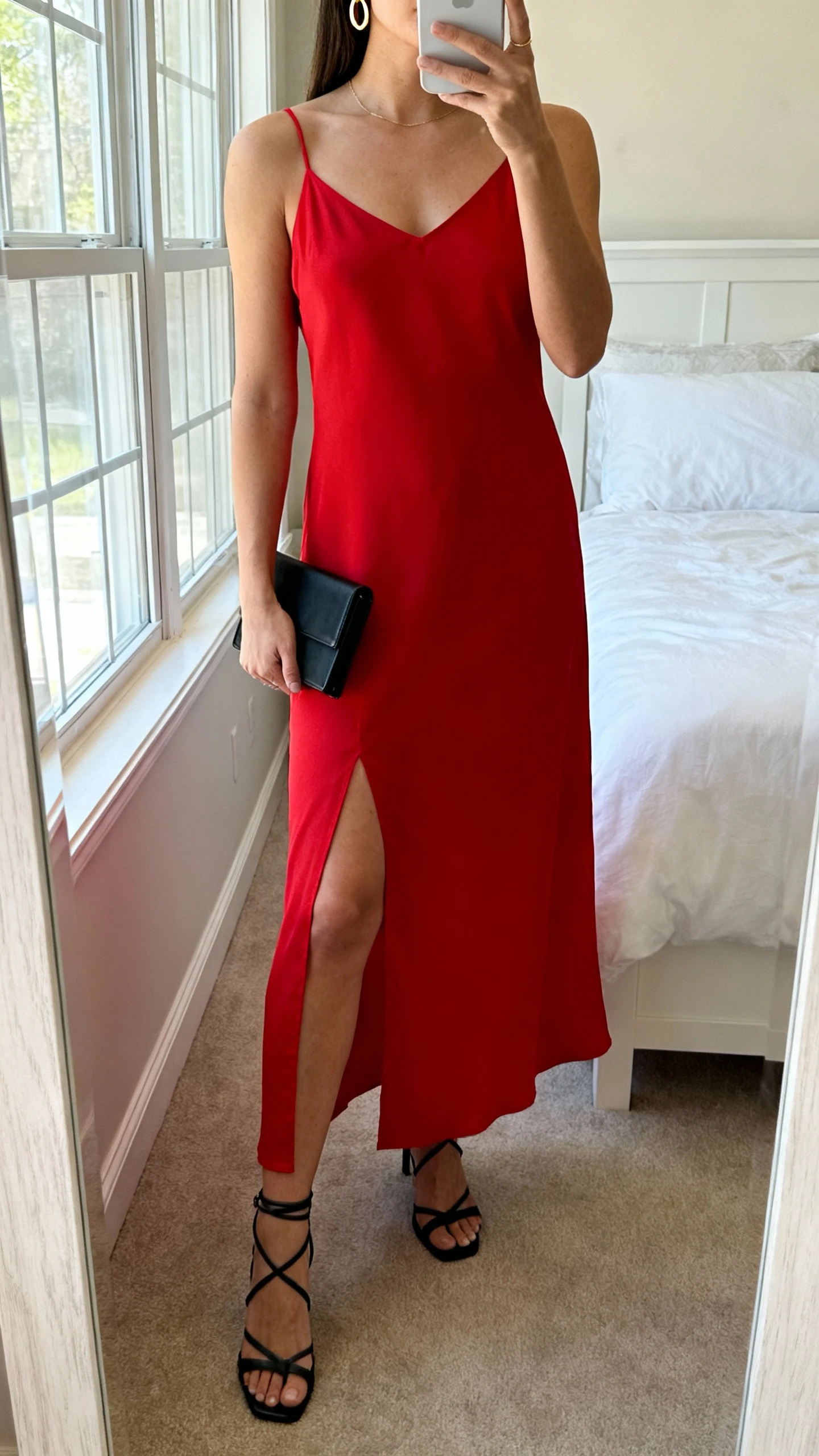 iPhone mirror selfie of a woman wearing a modern red slip dress with a thigh slit, delicate gold hoop earrings, strappy black heels, a slim black clutch, face not visible, cropped at neck, bedroom mirror, natural window light, iPhone photo quality.