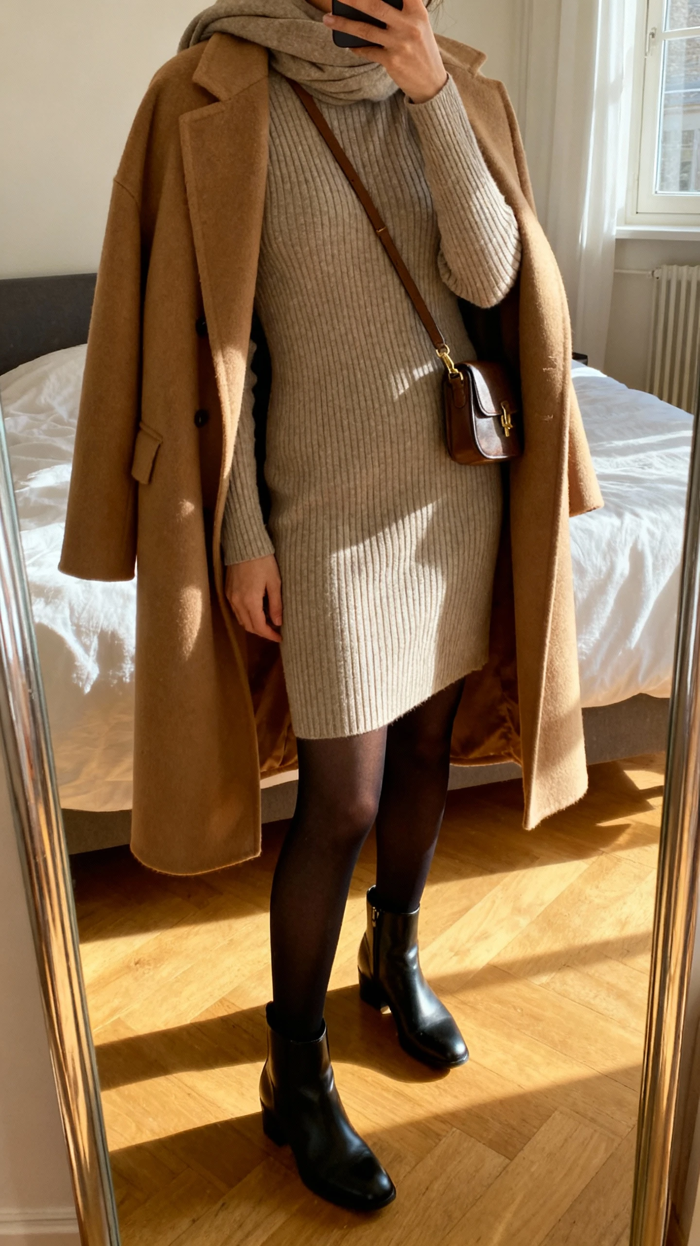 iPhone mirror selfie of a woman wearing a Parisian-style ribbed sweater dress, opaque black tights, sleek ankle boots, a camel wool coat draped over shoulders, small crossbody bag, and a simple scarf, face not visible, cropped at neck, bedroom mirror with natural morning window light, iPhone photo quality.