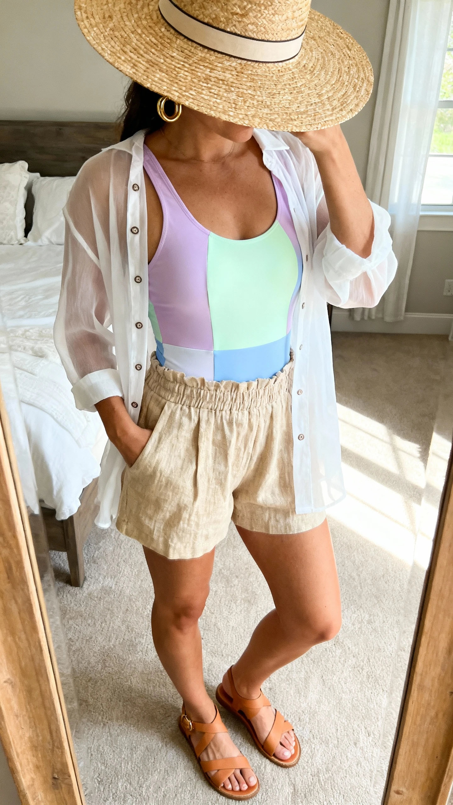 iPhone mirror selfie of a woman wearing a pastel scoop-neck one-piece swimsuit (lavender/pistachio/powder blue) with a sheer white button-up cover-up, high-waisted linen shorts in sand, tan strappy flat sandals, an oversized straw hat with a slim ribbon, and gold hoops; face not visible, cropped at neck, bedroom mirror, soft morning window light, iPhone photo quality.