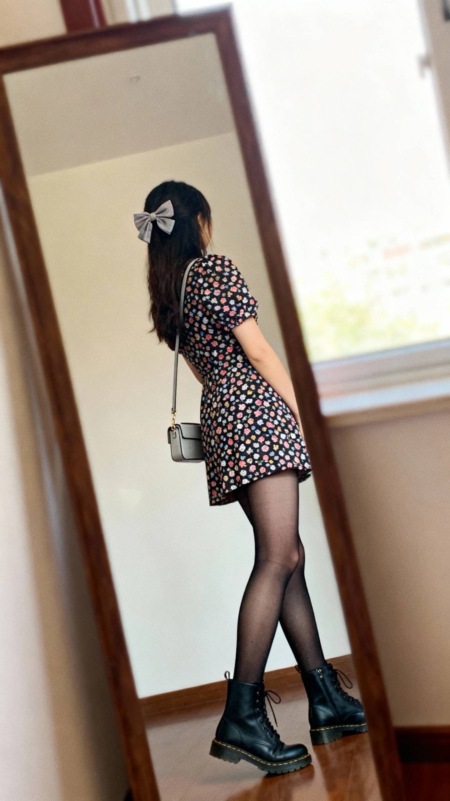 iPhone mirror selfie of a woman wearing a playful mini dress with sheer black tights, ankle boots, and a hair bow, carrying a small crossbody, face not visible, angled side view in a full-length mirror by a window with natural light, iPhone photo quality.